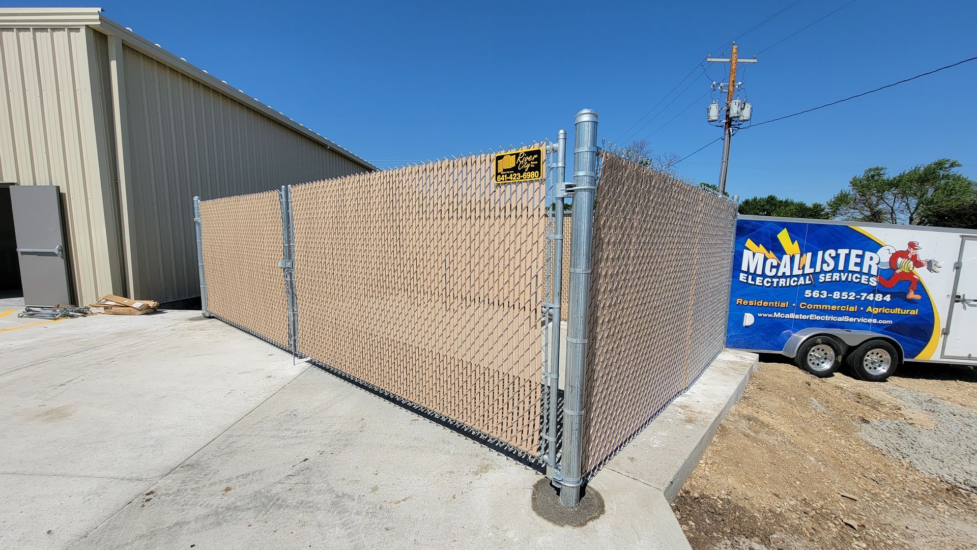 Chain-link gate beside a beige warehouse, with a blue utility trailer in a sunny lot