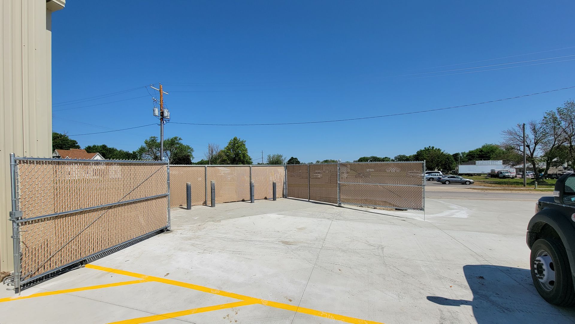 Sunlit empty parking lot beside a wooden fence and tan building under a clear blue sky