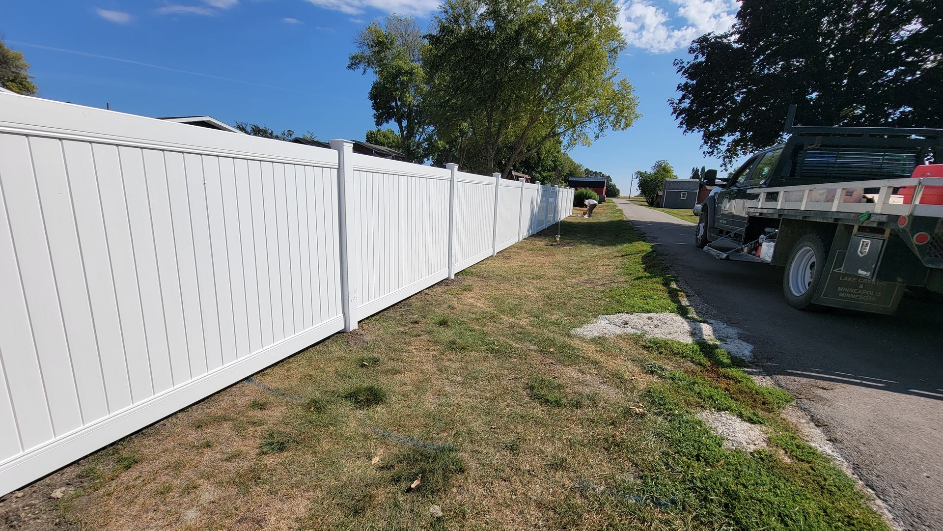 White vinyl fence beside a grassy roadside, with a truck parked on the right under a blue sky.