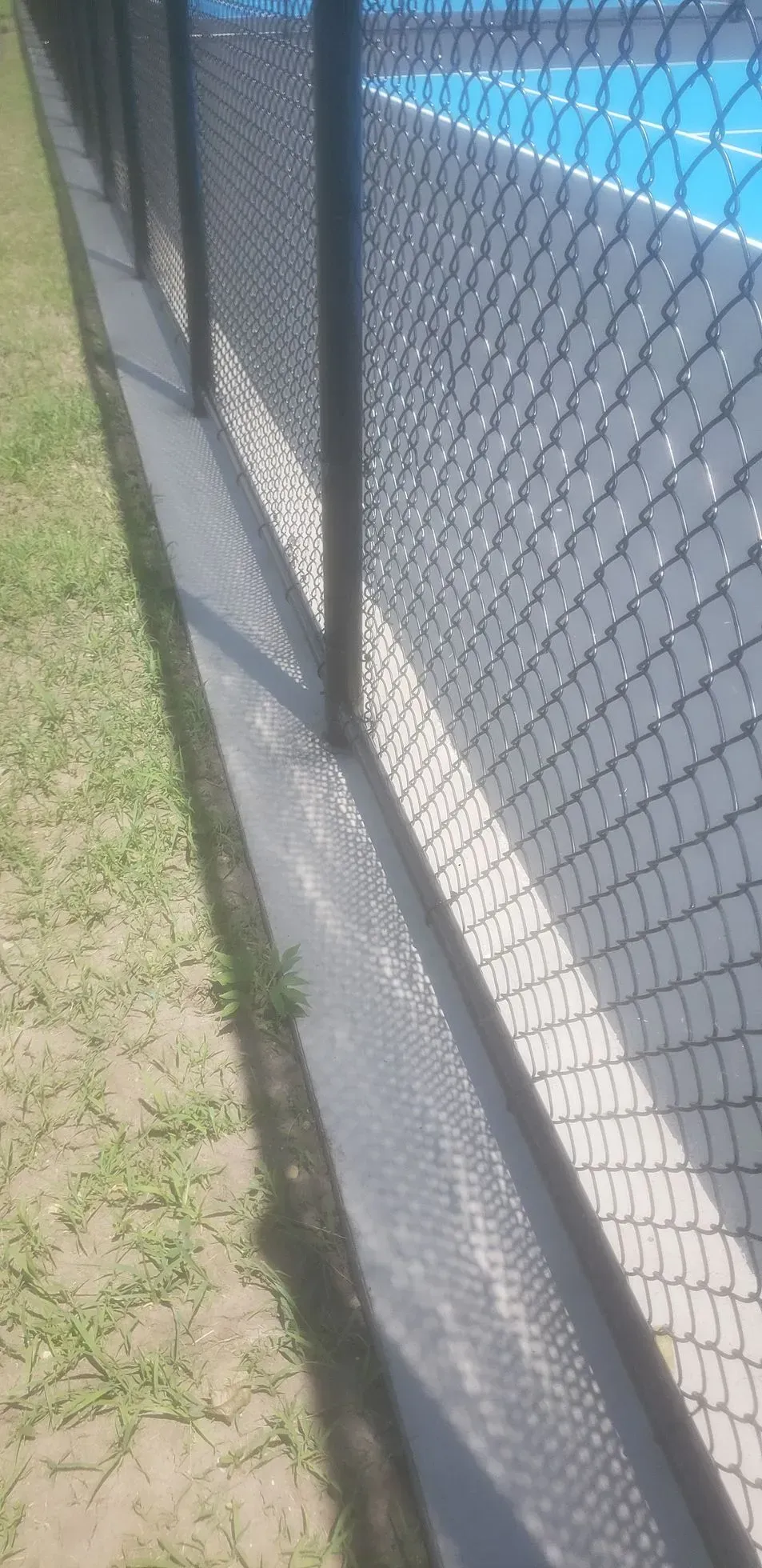 Chain-link fence beside a blue tennis court with grass along the edge