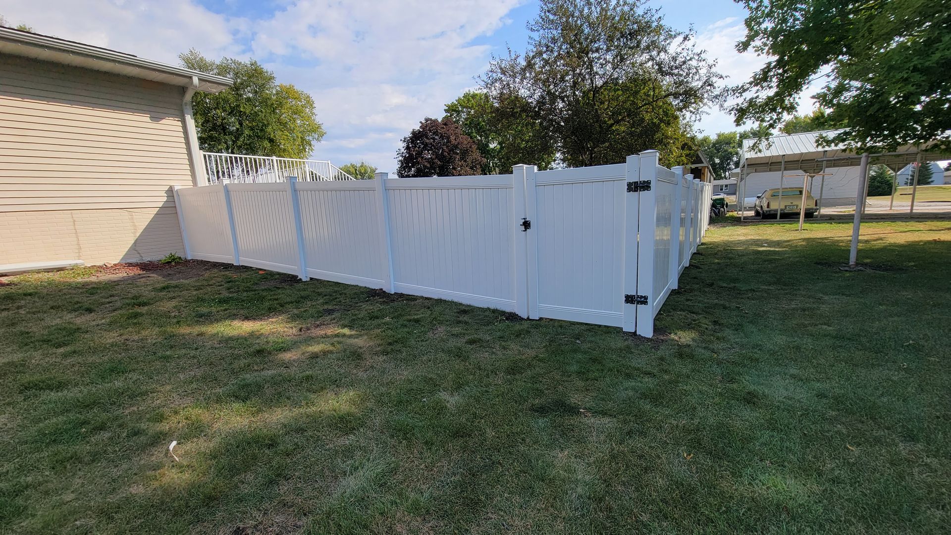 White vinyl fence panels leaning along a grassy yard beside a beige house under a partly cloudy sky