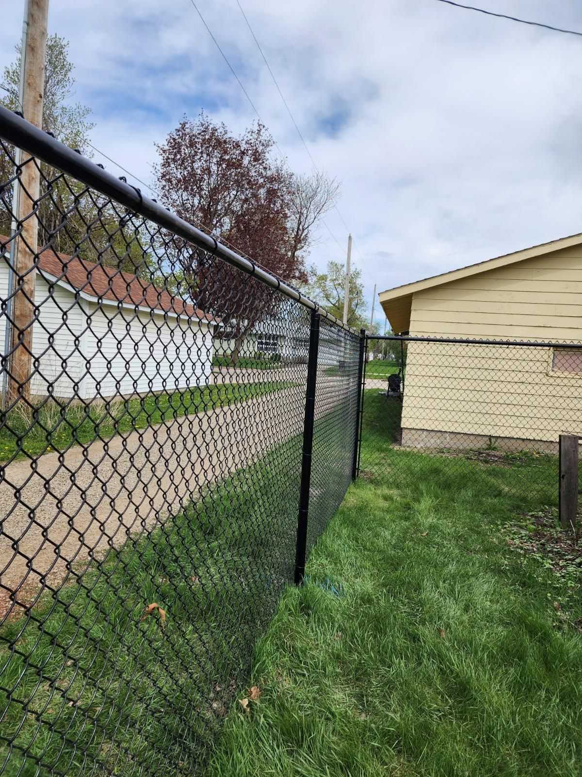 Chain-link fence beside a gravel driveway and beige house, with green grass and cloudy sky.
