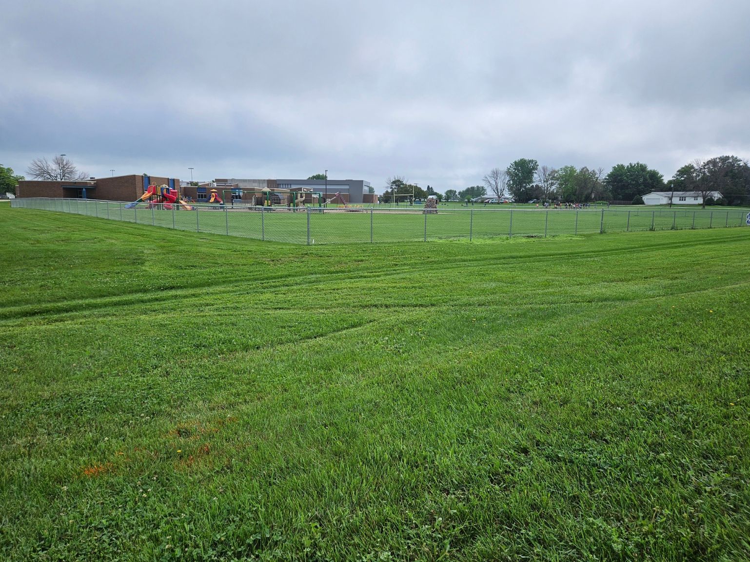 Grassy soccer field with a chain-link fence, overcast sky, and people gathered near buildings in the distance