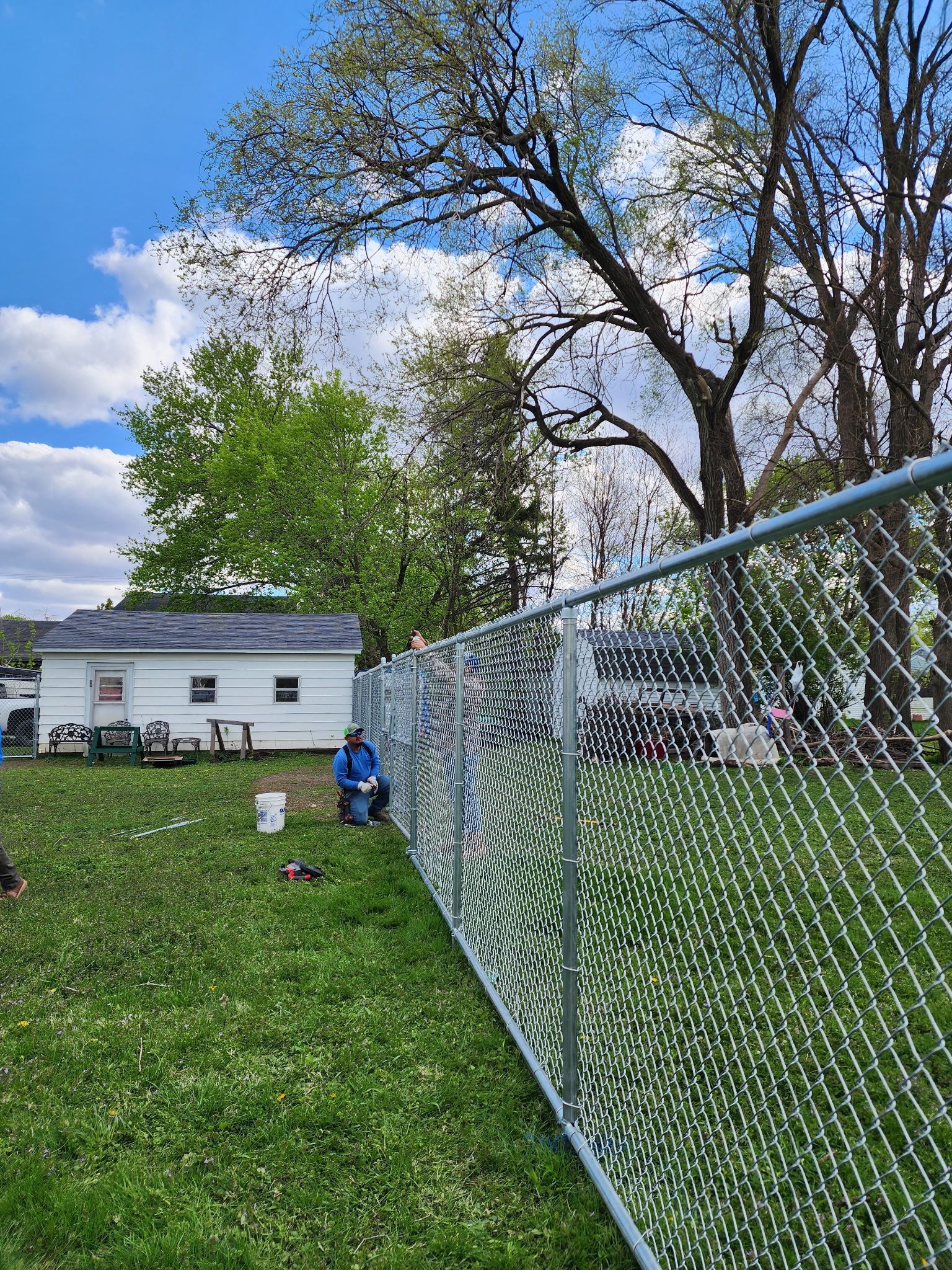 Chain-link fence in a grassy yard with a white house, trees, and a person in blue sitting outside under a cloudy sky