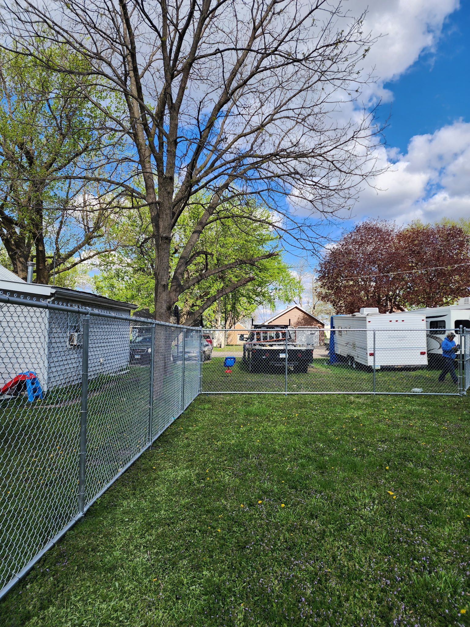 Backyard with chain-link fence, green lawn, trees, and a patio table under a blue sky.