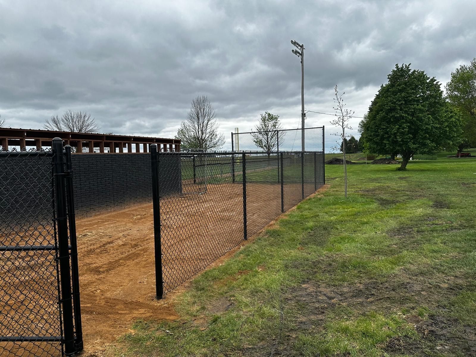 Chain-link fence beside a dirt baseball field under a cloudy sky, with trees and a light pole in the background