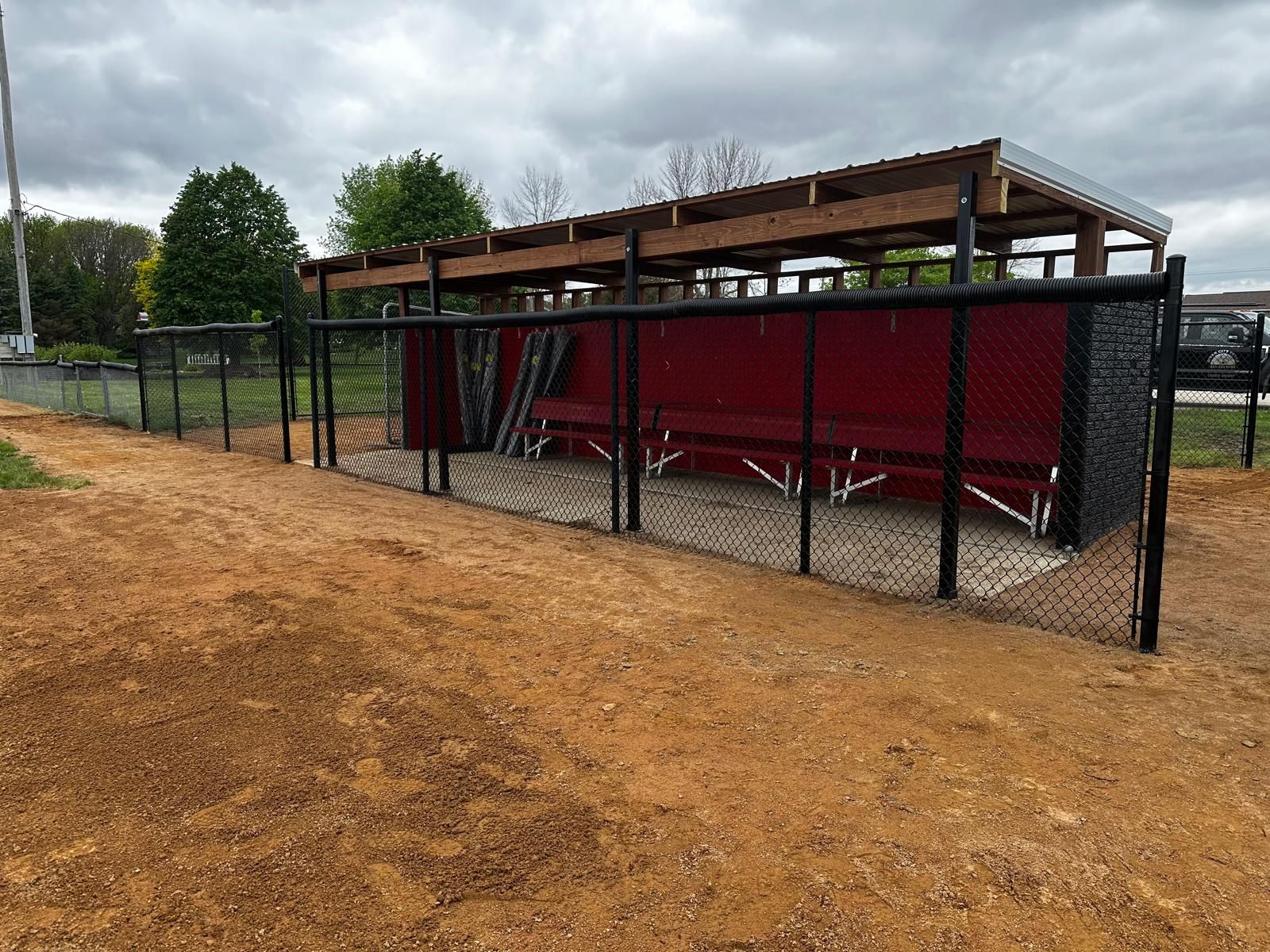 Chain-link fenced outdoor shelter with a red roof and seating on a dirt lot under cloudy skies