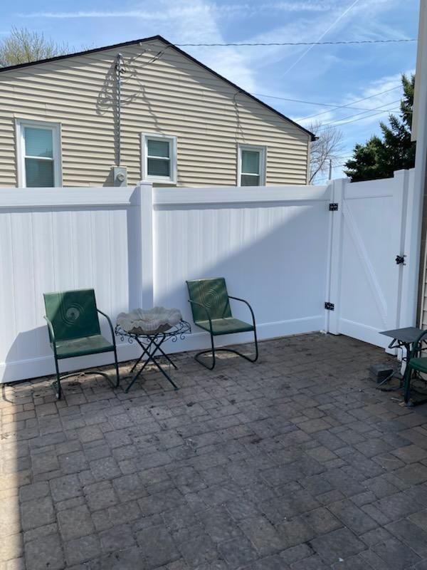 Small fenced patio with two green chairs and a table on stone pavers, backed by a white privacy fence
