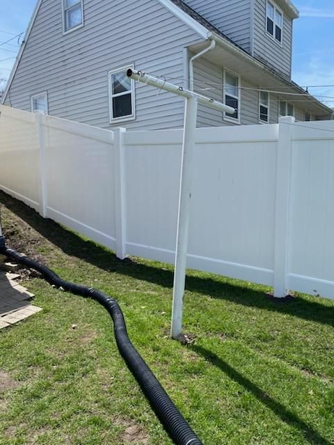 White vinyl fence beside a gray house with a black drainage hose on the grass