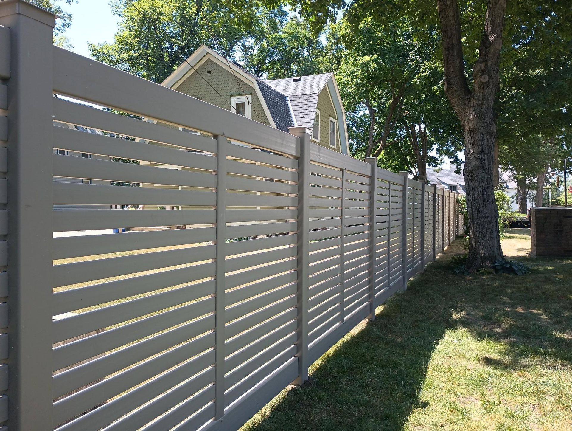 Long white slatted fence along a grassy yard beside trees and houses on a sunny day