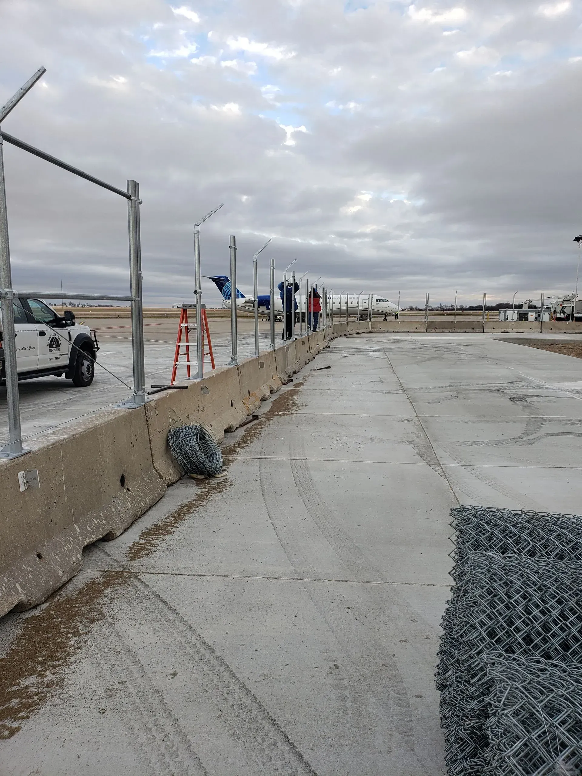 Concrete barrier along a cracked roadside with construction fencing, ladders, and workers under a cloudy sky