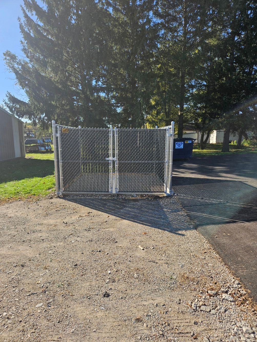 White metal cage enclosure by a tree-lined gravel lot, with a blue bin in the background.
