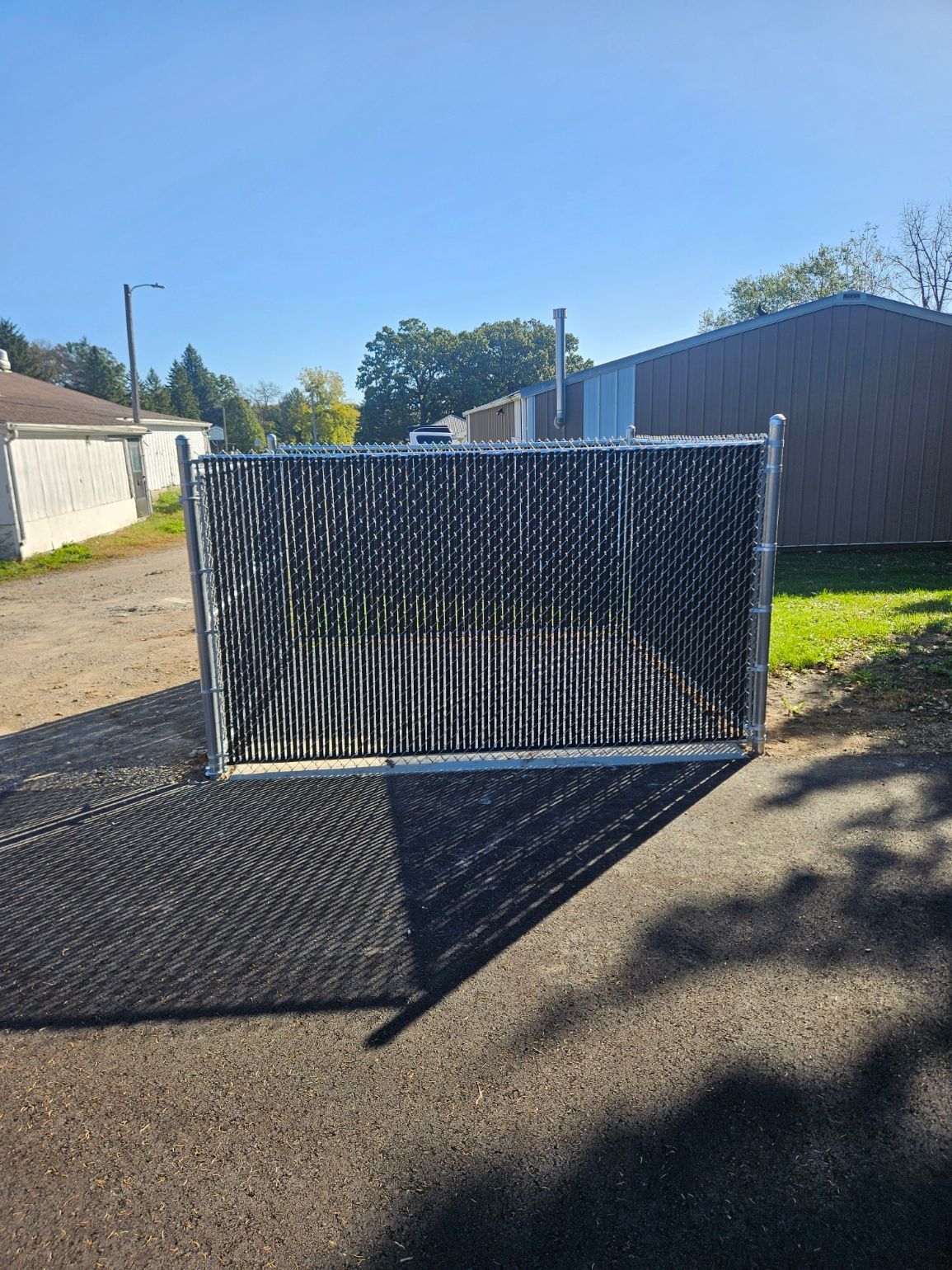 Black metal gate across a gravel driveway in front of a house, casting a long shadow in sunlight.