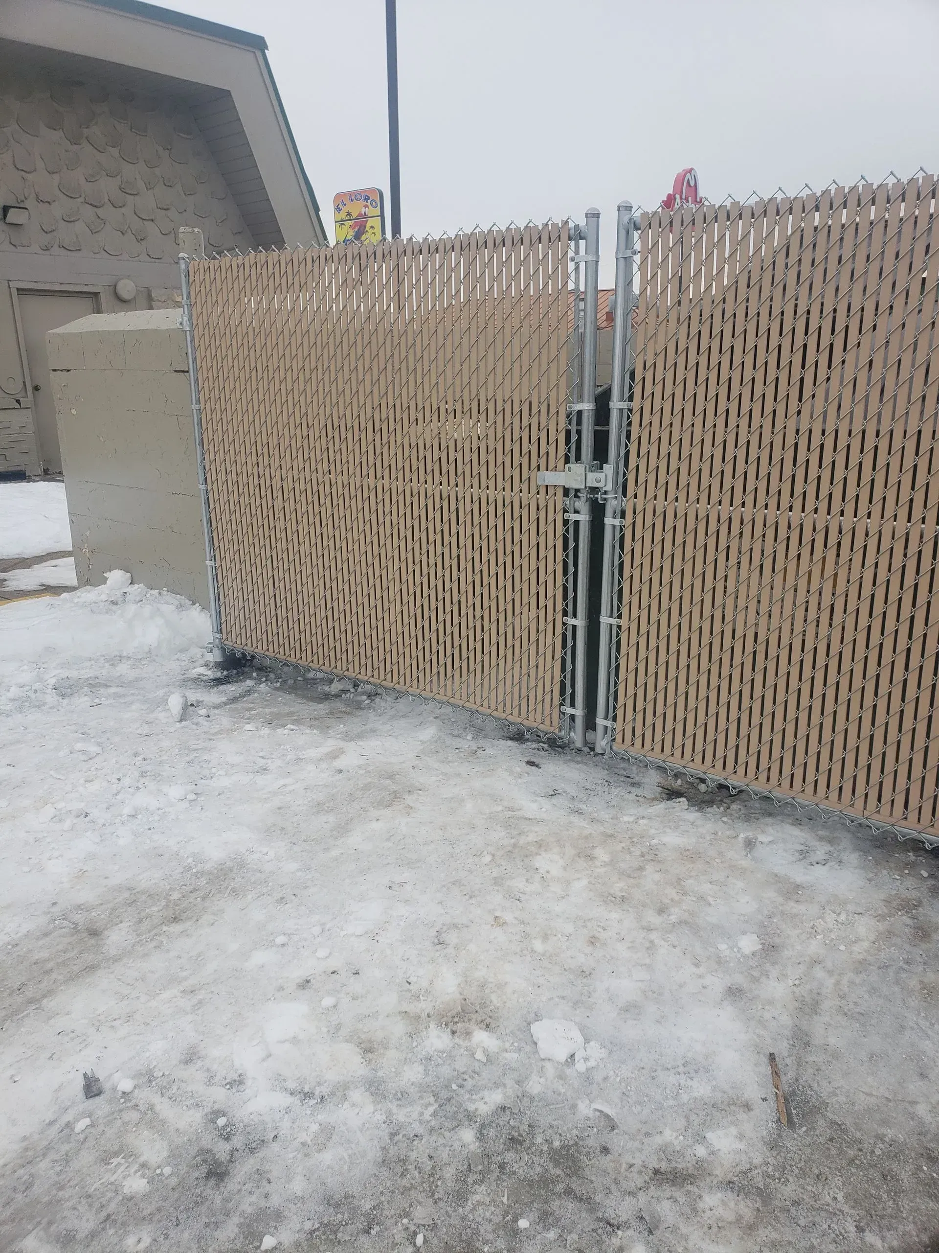 Snowy driveway with a tan woven gate beside a gray building