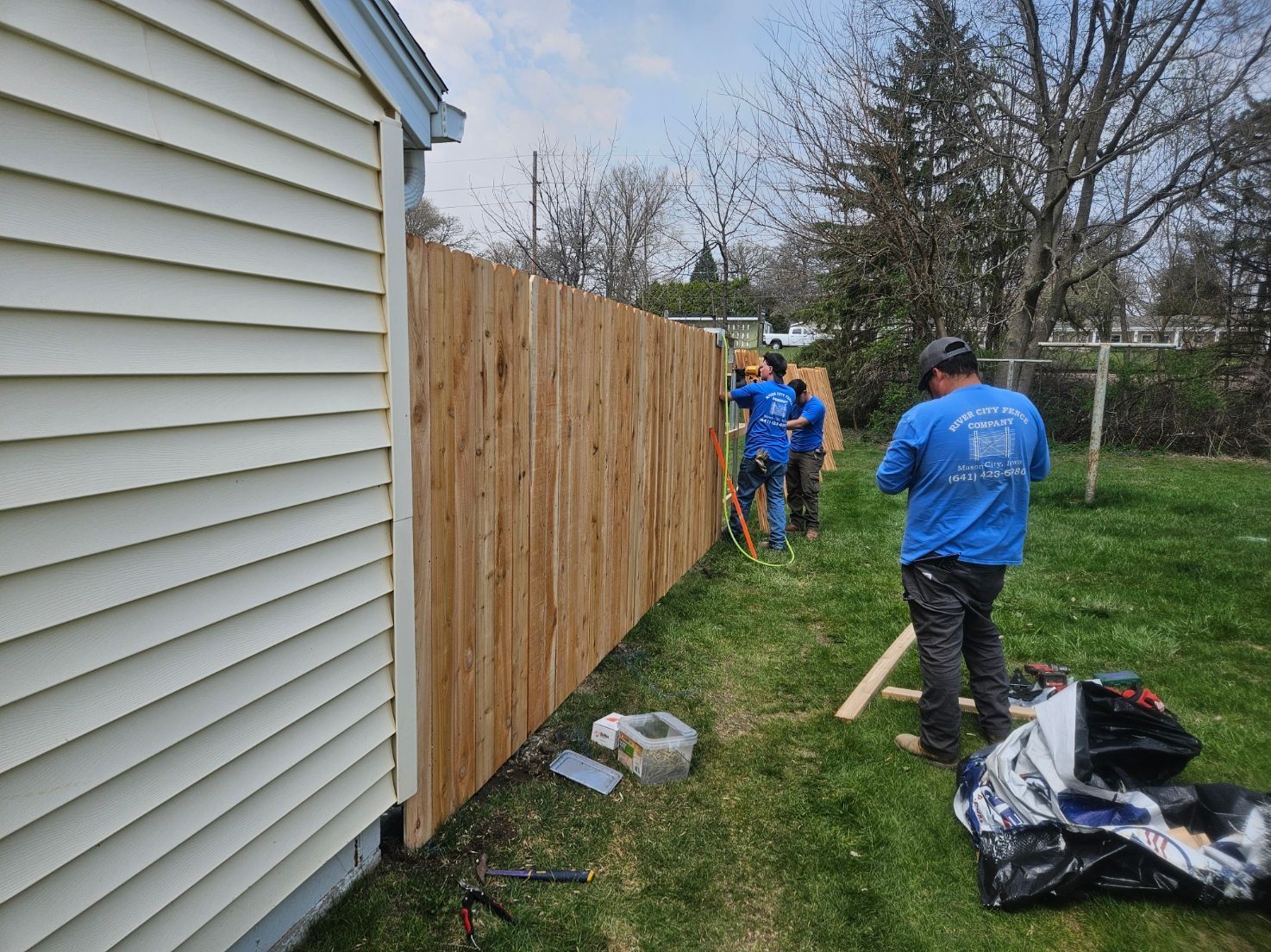 Workers in blue shirts install a wooden fence beside a house on a grassy yard.