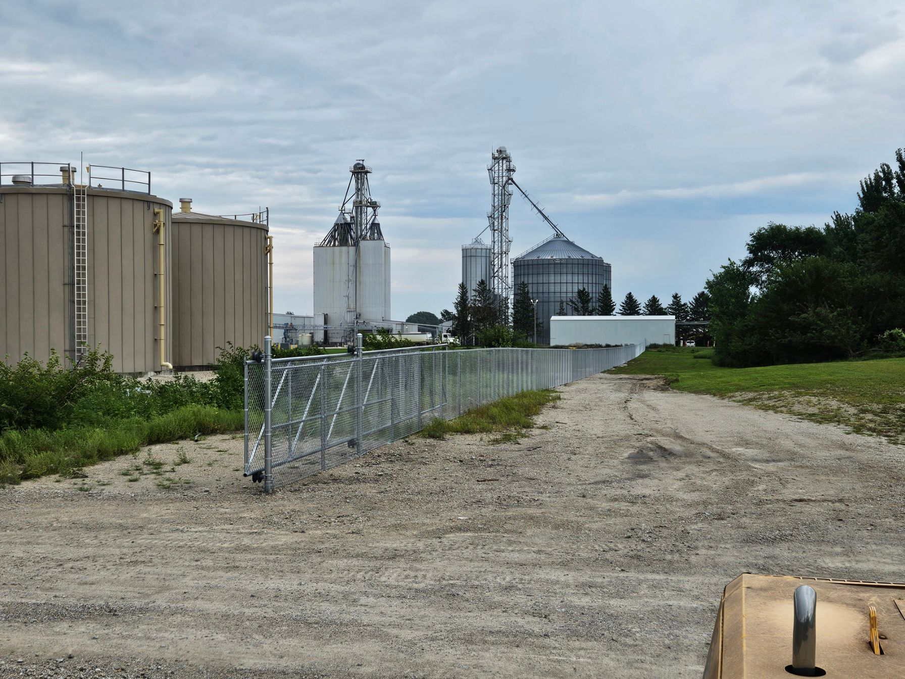 Gravel road beside grain silos and metal fencing at an industrial facility under a cloudy sky