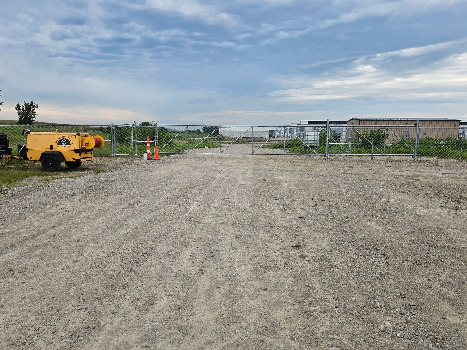 Gravel construction site with a yellow generator, orange cones, and a fenced building under a cloudy sky