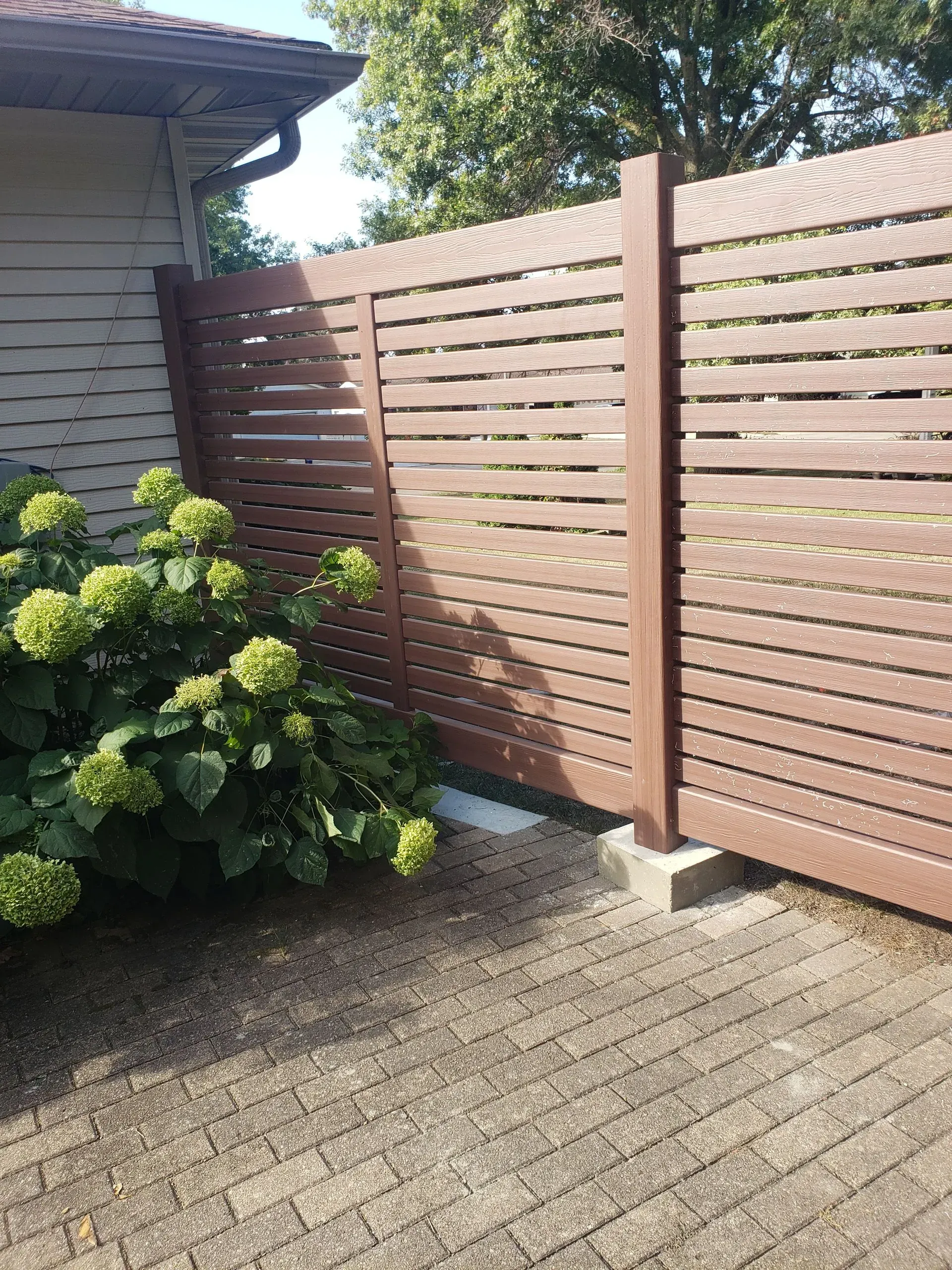 Wooden slat fence beside a house, with a leafy shrub and paved patio in sunlight