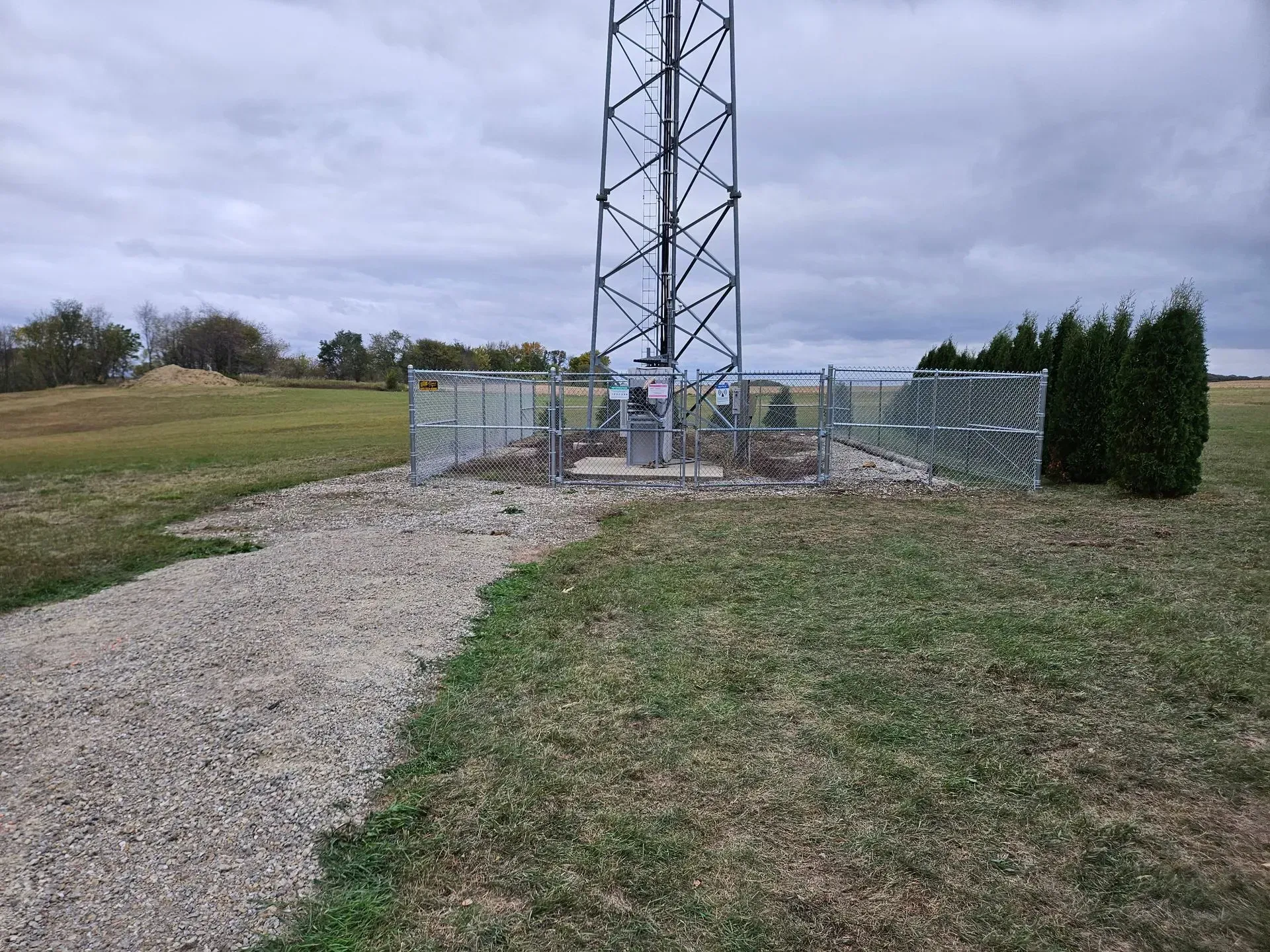 Gravel path leading to a fenced utility tower on a grassy hill under cloudy skies