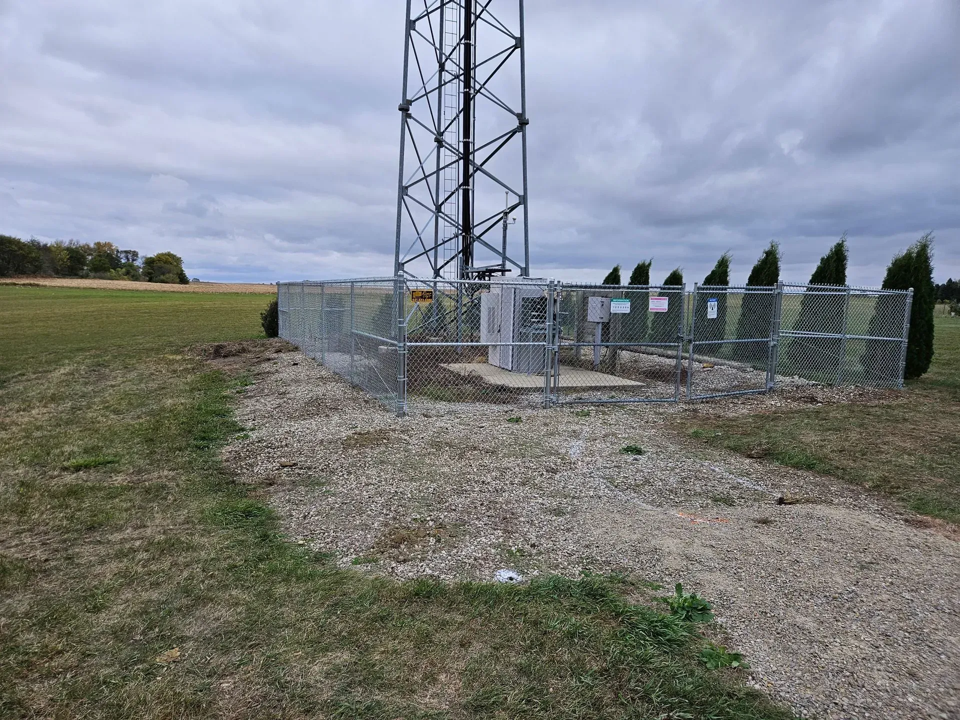 Fenced utility tower on a gravel pad beside a grassy field under cloudy sky