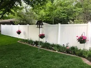 White vinyl fence with hanging pink flowers along a green lawn and garden bed