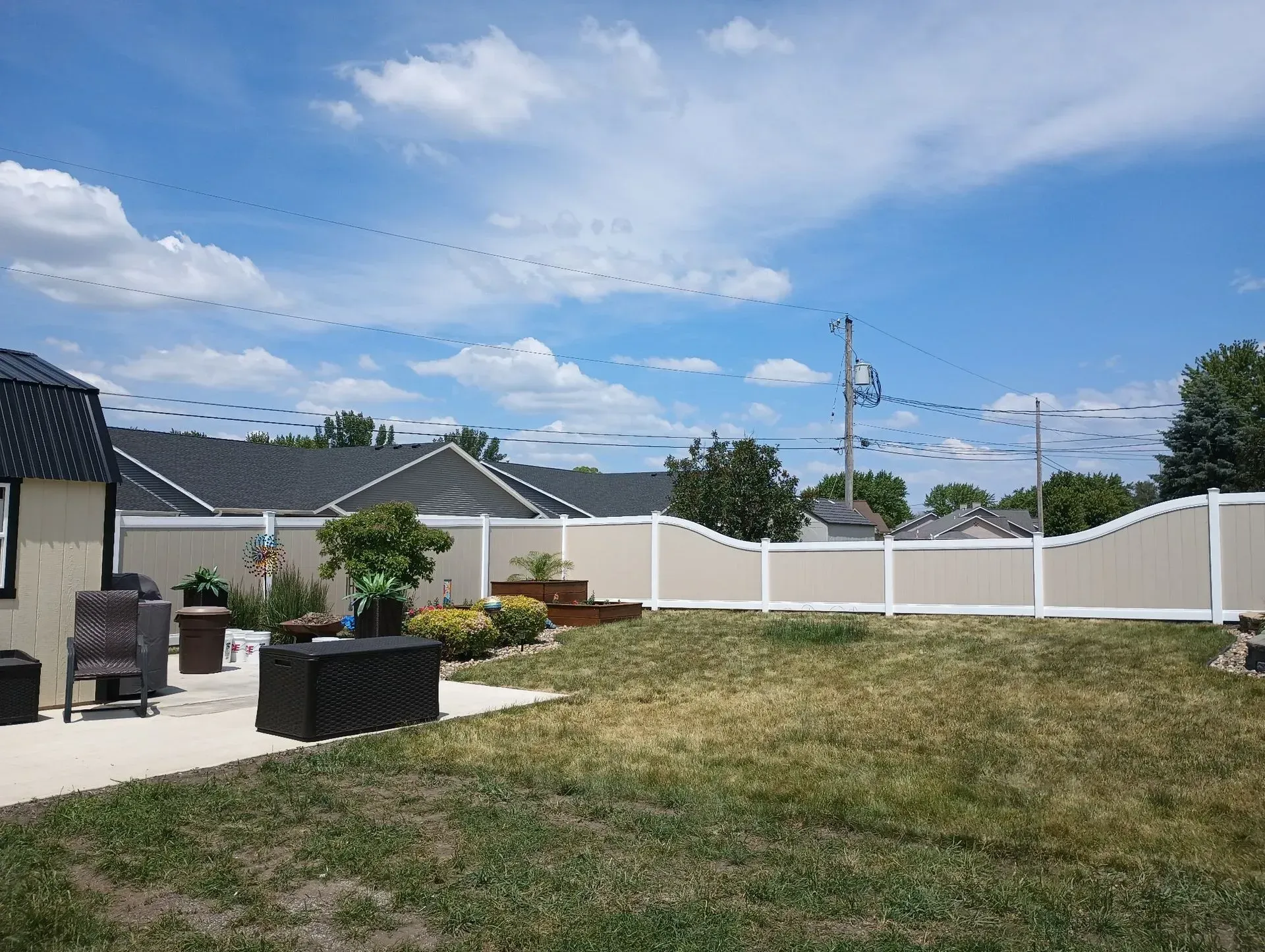 Backyard with grassy lawn, patio seating, white fence, and blue sky with clouds
