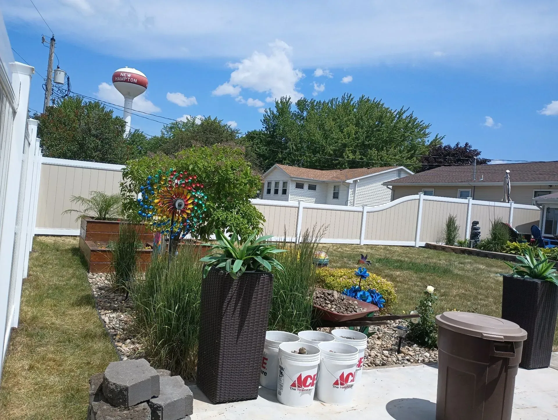 Backyard garden with white fence, potted plants, lawn, and a water tower in the distance.