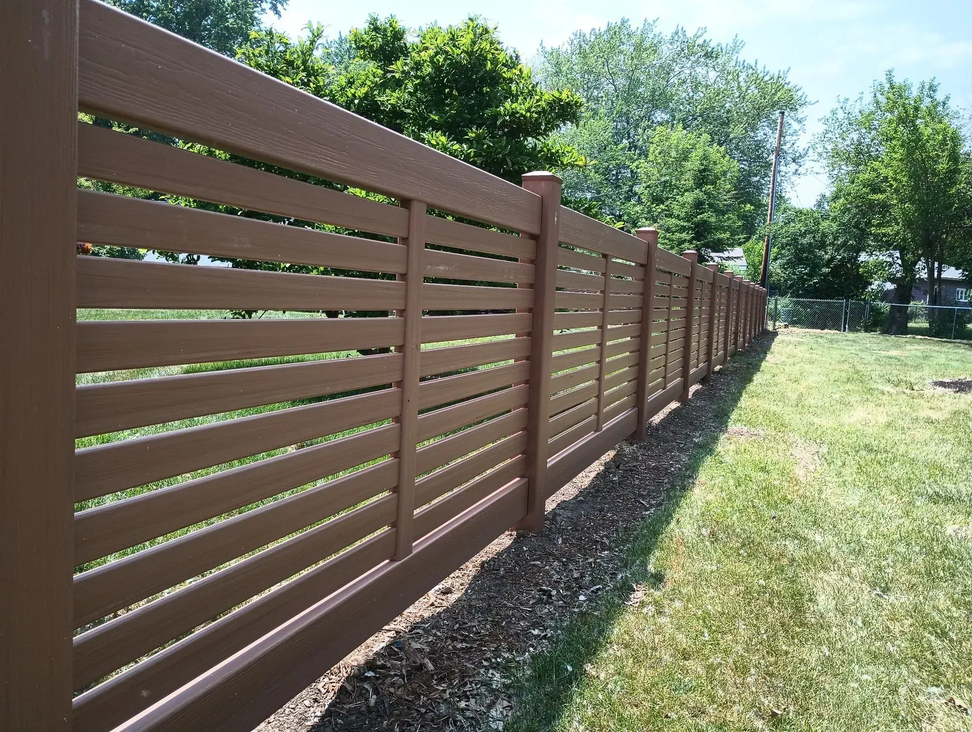 Brown wooden fence lining a sunny grassy yard with trees in the background