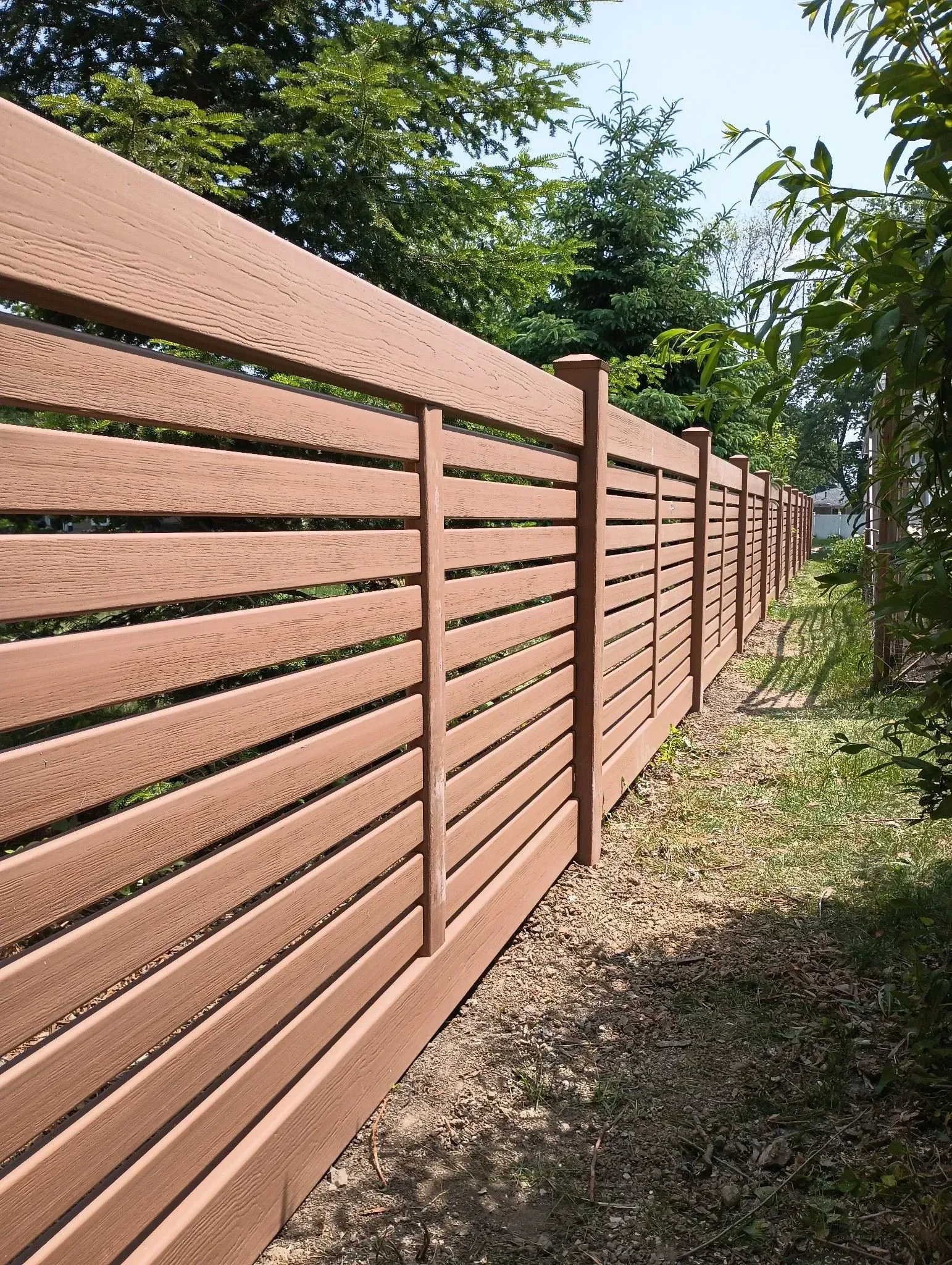 Brown wooden privacy fence with horizontal slats along a grassy path beside trees