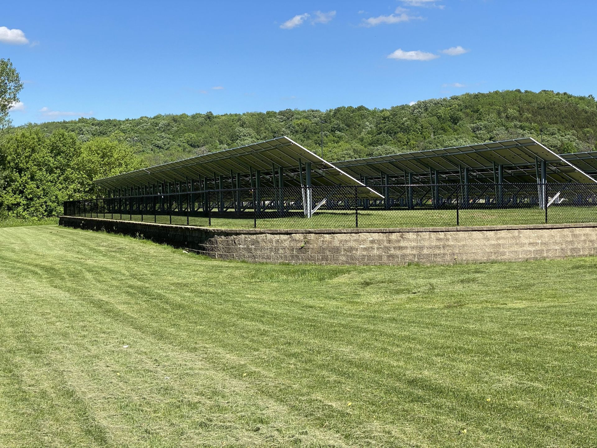 Solar panels on a stone wall in a grassy field, with green hills and blue sky behind.