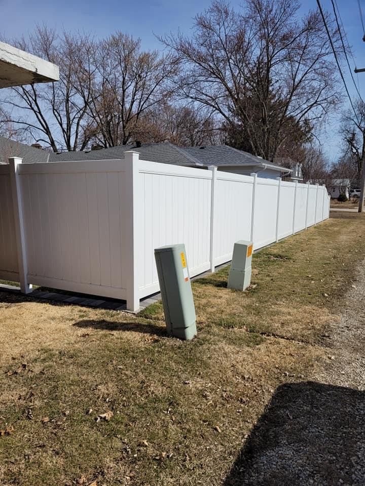 White vinyl privacy fence along a grassy roadside with utility boxes in front and bare trees behind