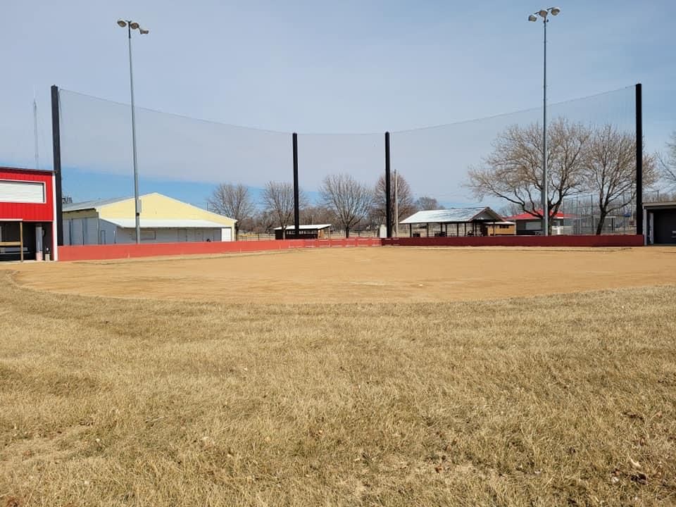 Empty baseball field with brown grass, red dirt infield, and tall lights under a clear blue sky.