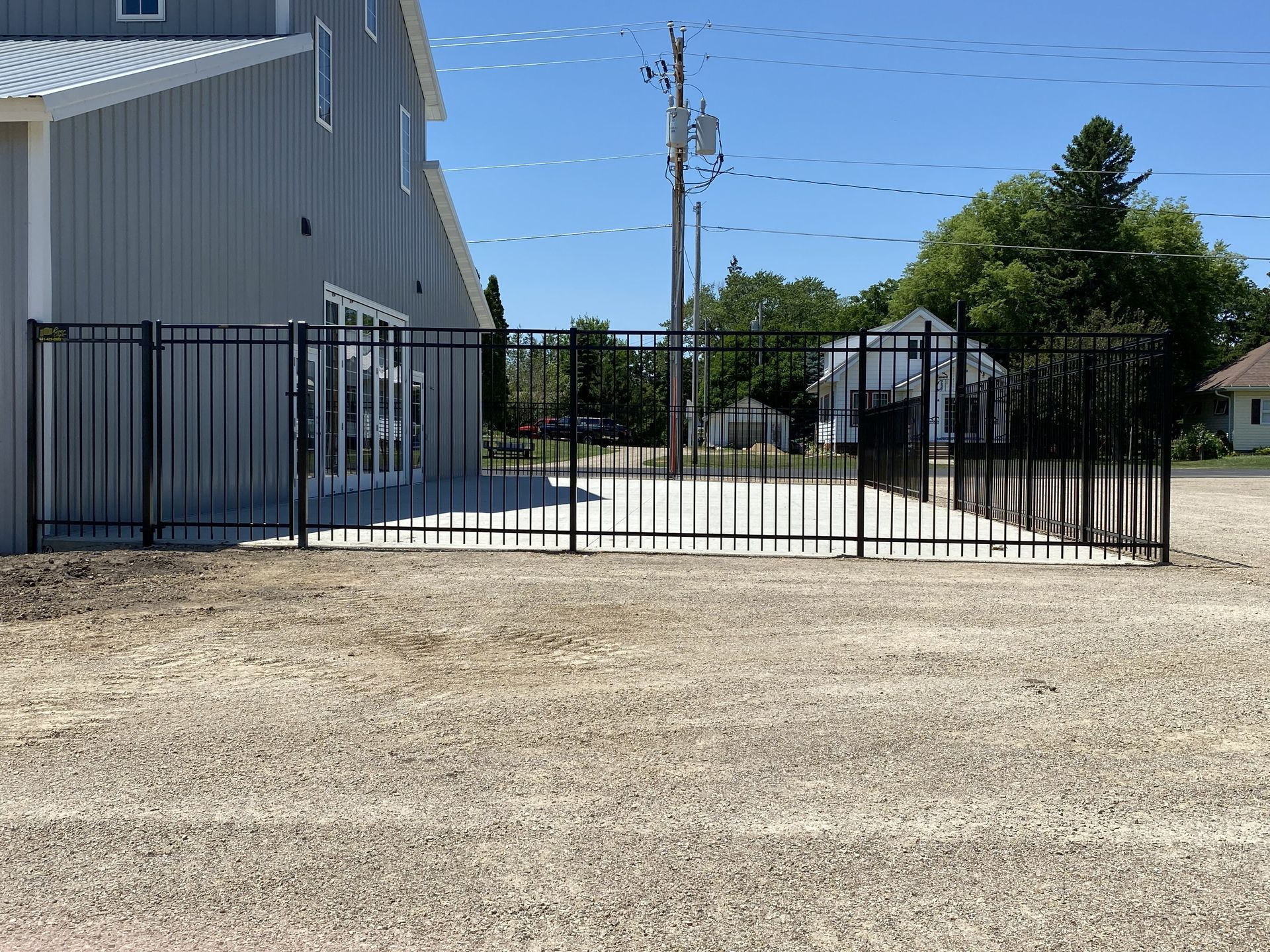 Black metal gate across a gravel driveway beside gray industrial buildings under a blue sky.