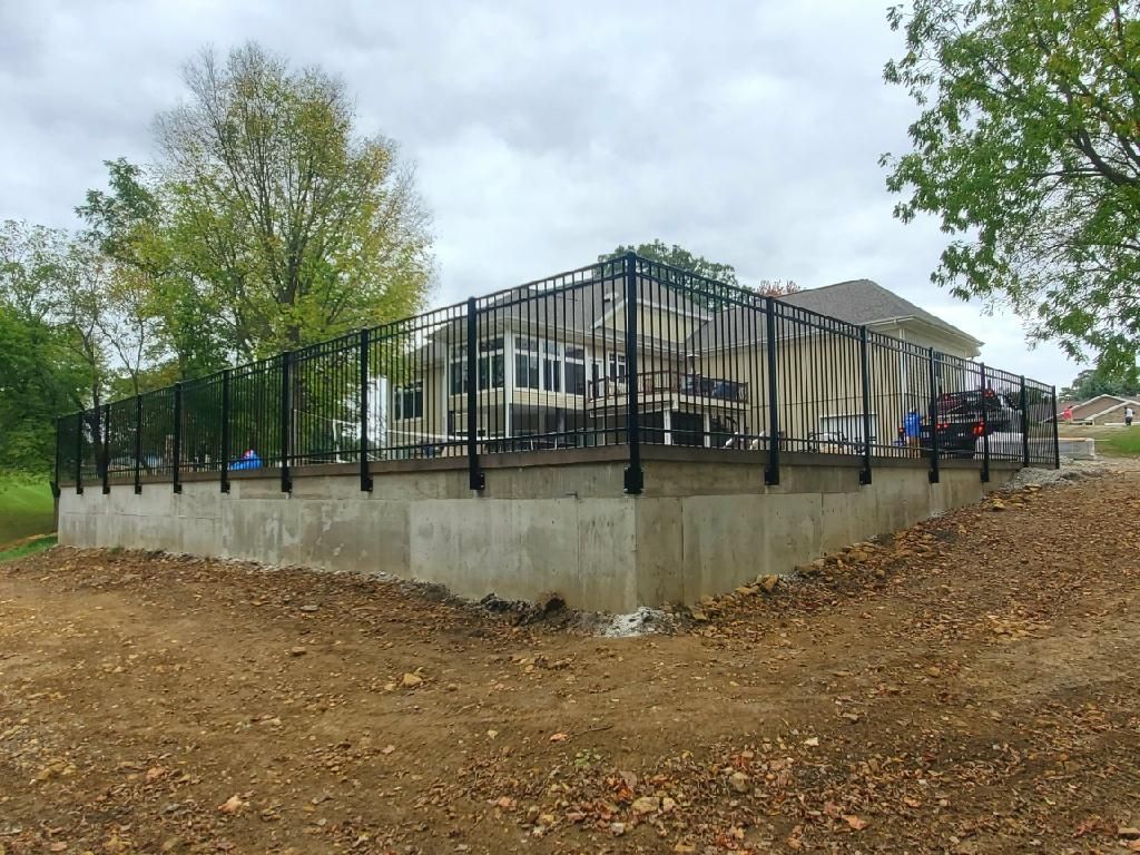 Concrete building with black metal railings on a raised lot, surrounded by trees and dirt ground