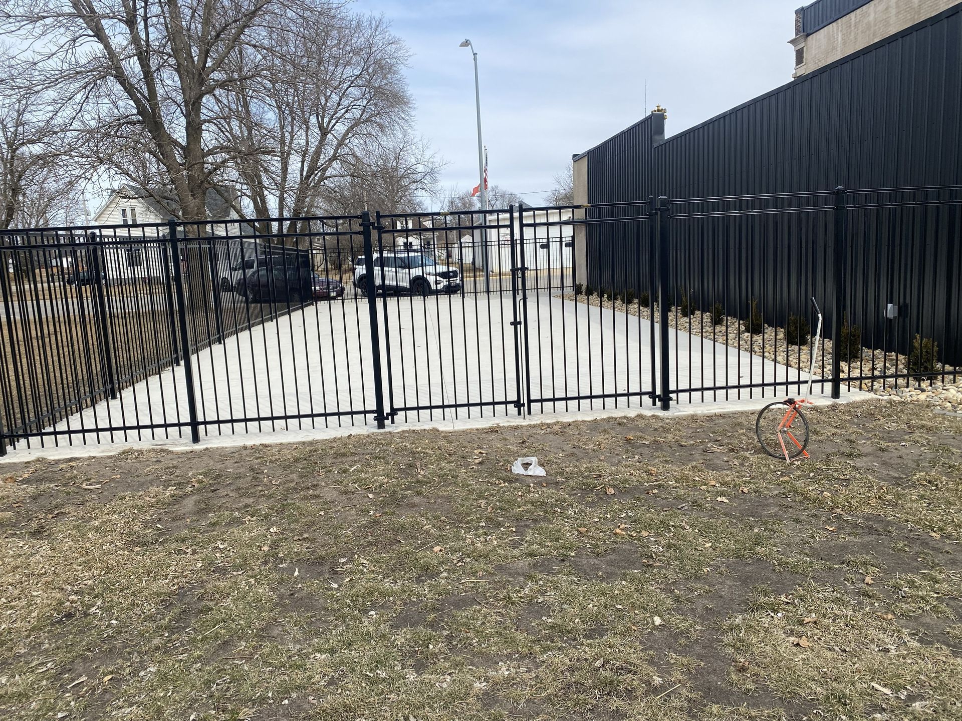 Black metal fence across a vacant lot with leafless trees and a building in the background