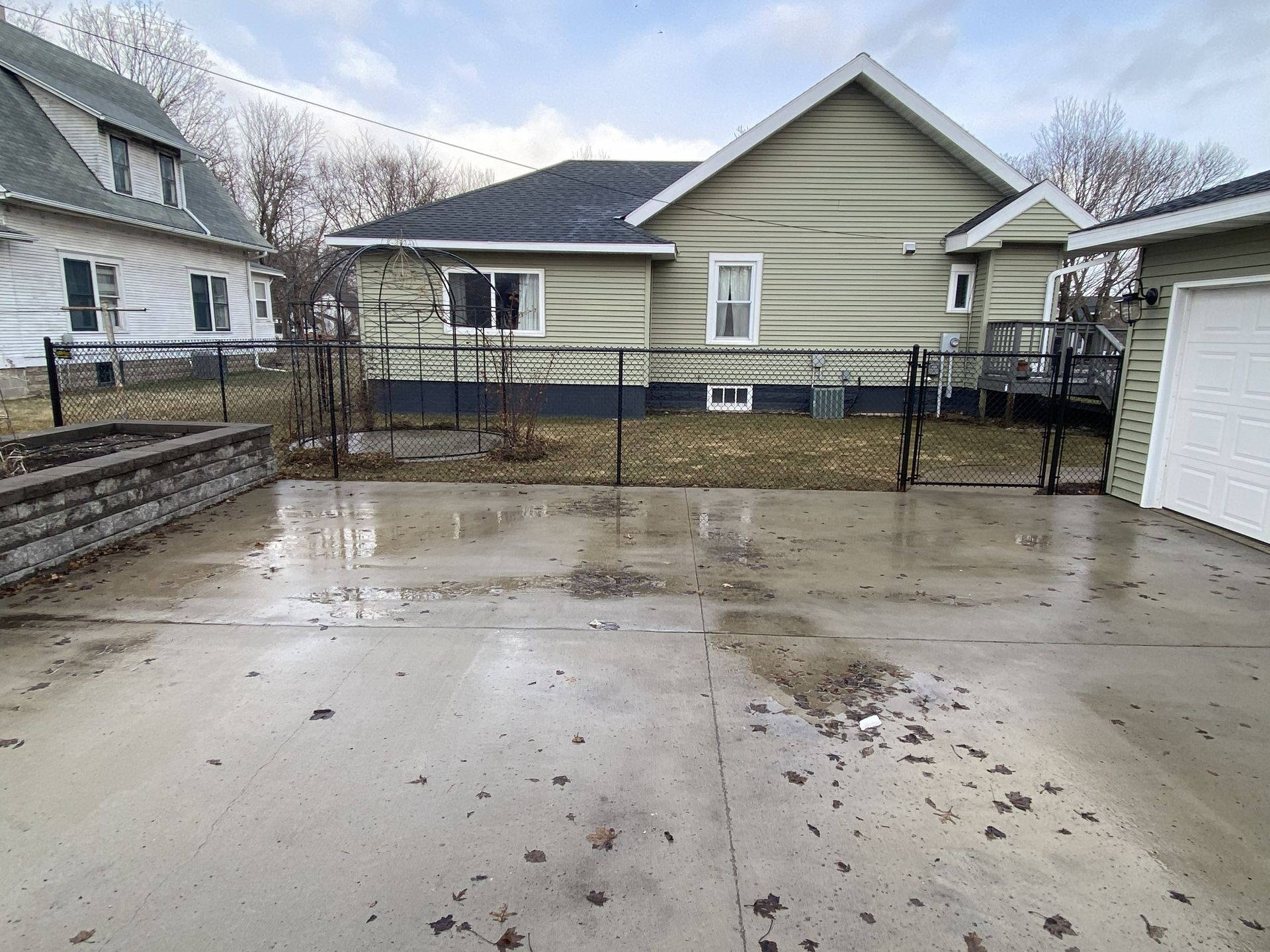 Wet concrete driveway and fenced backyard behind a pale yellow house on a cloudy day