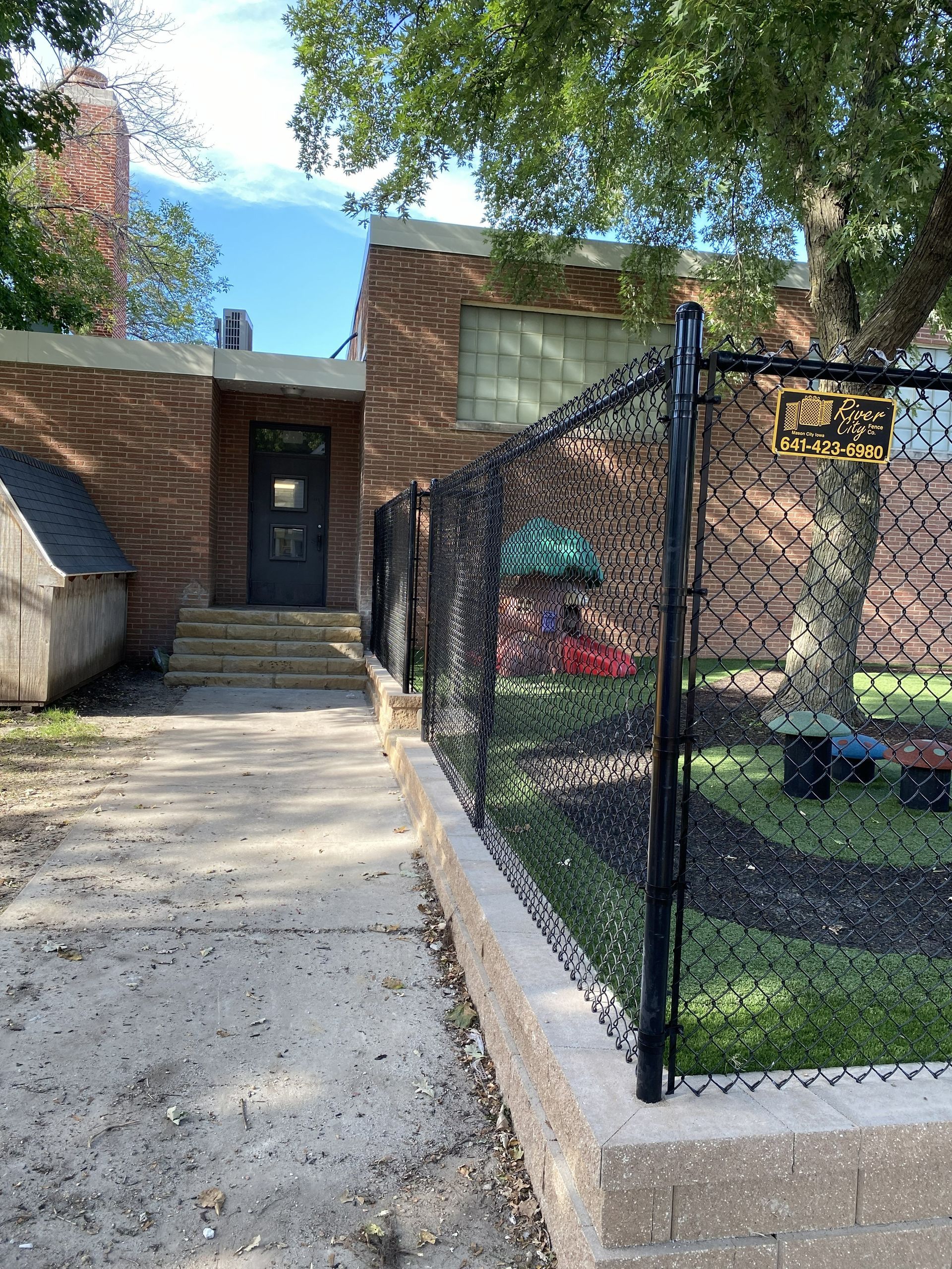 Concrete walkway leading to a brick house with steps, black chain-link fence, and trees overhead