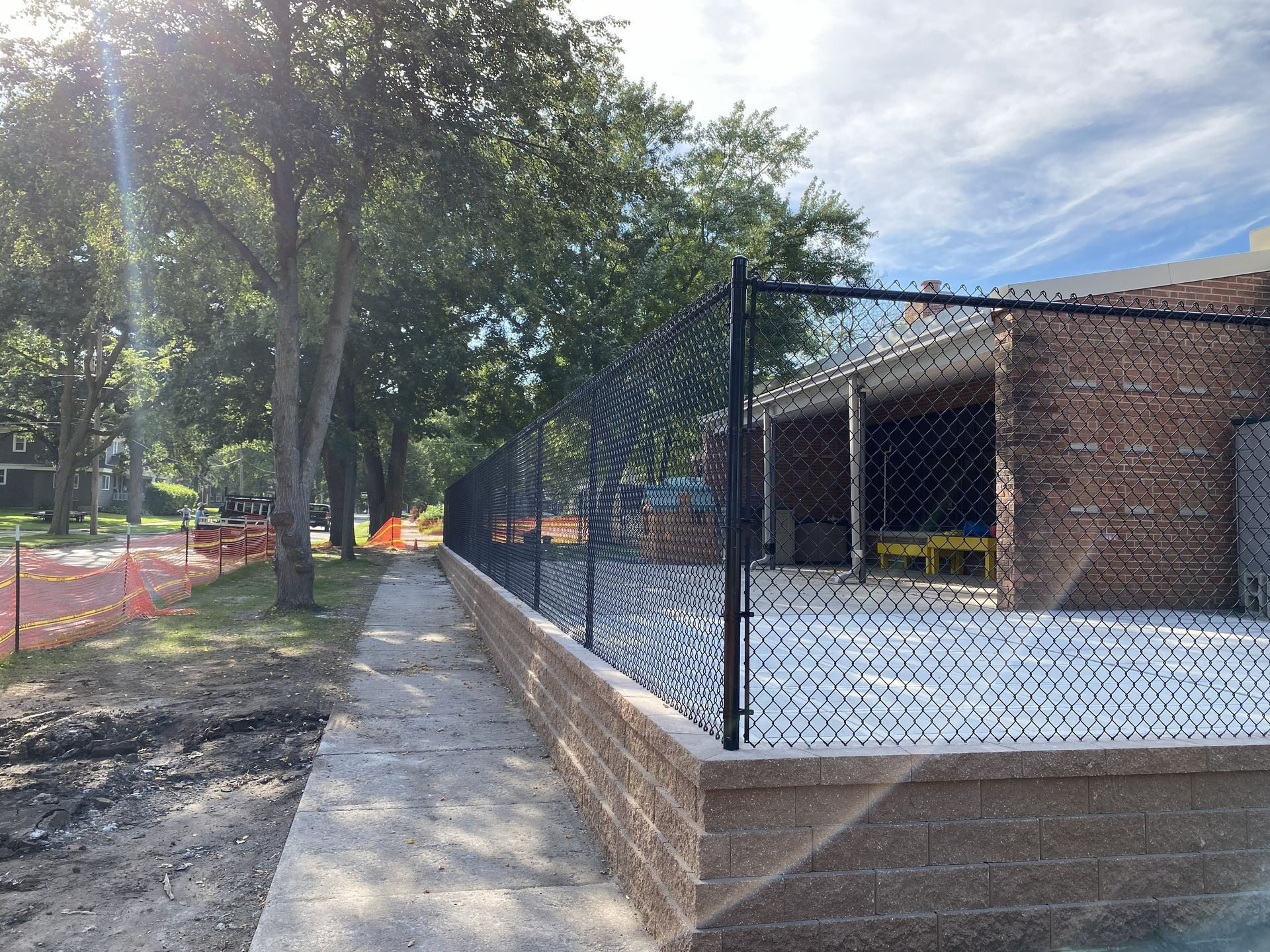 Chain-link fence beside a brick building along a tree-lined path on a sunny day