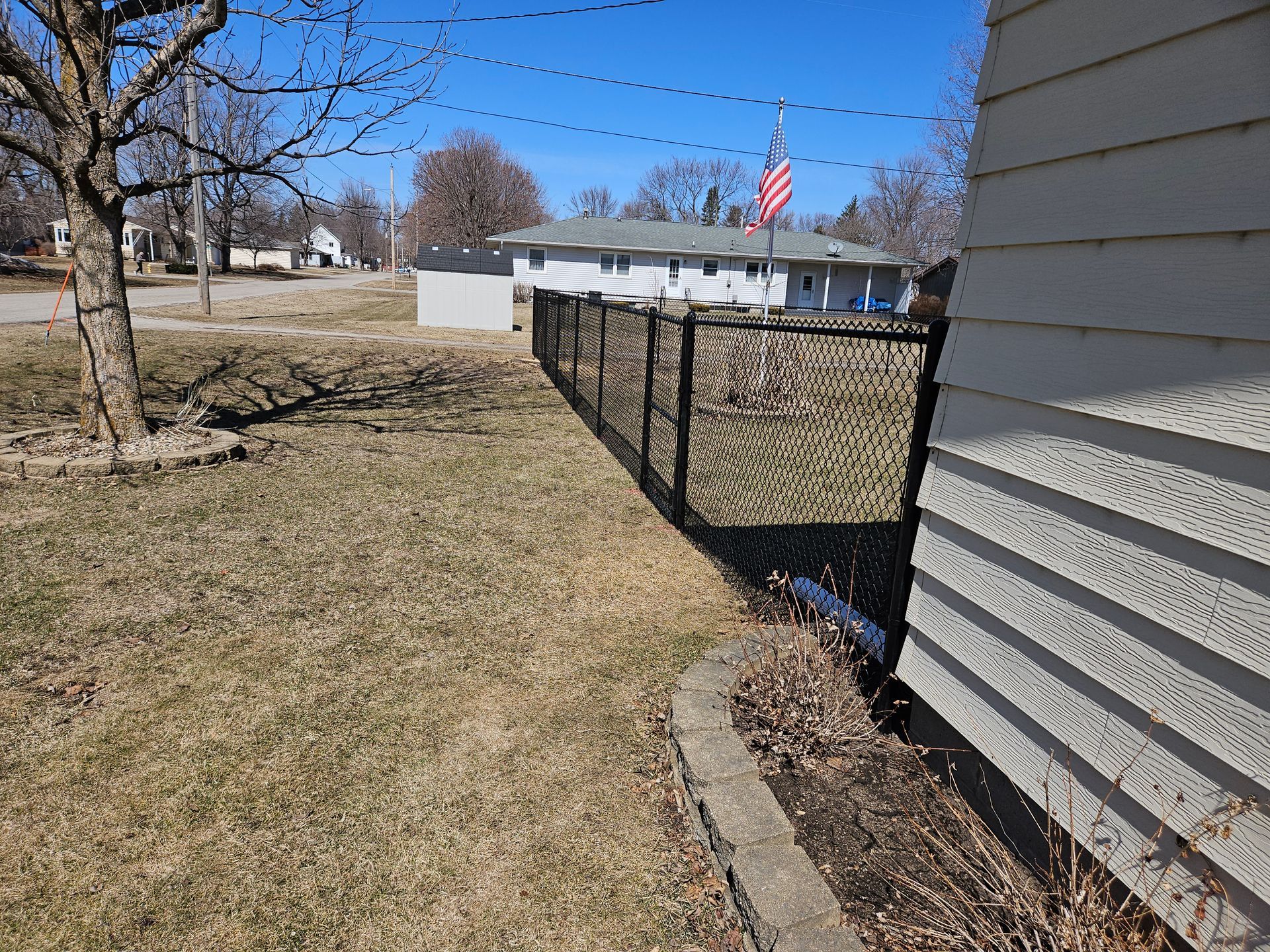 Suburban yard with chain-link fence beside a house, patchy grass, trees, and an American flag in the distance