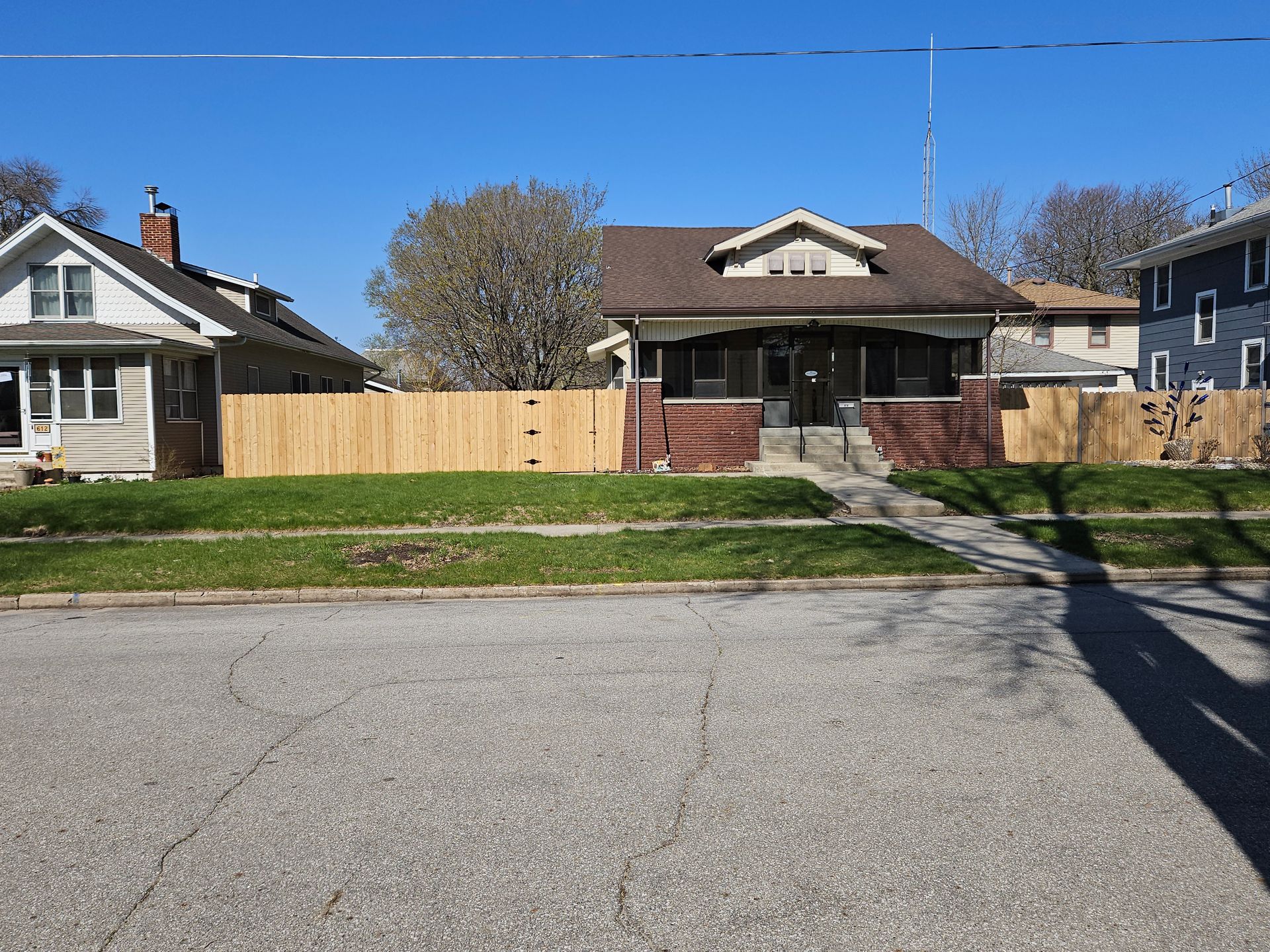 Quiet suburban street with two houses, a wooden fence, and a grassy front yard under clear skies.