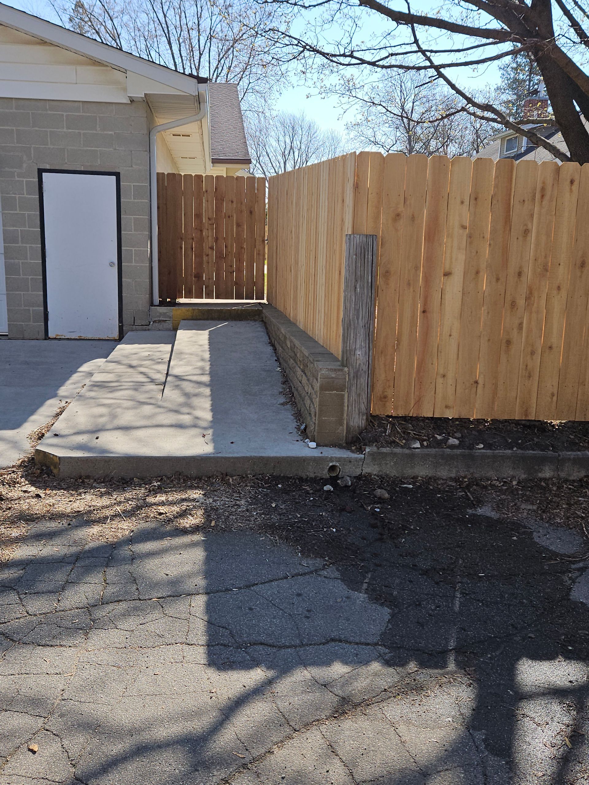 Concrete walkway beside a house, ending at a wooden gate and privacy fence in a sunny yard.