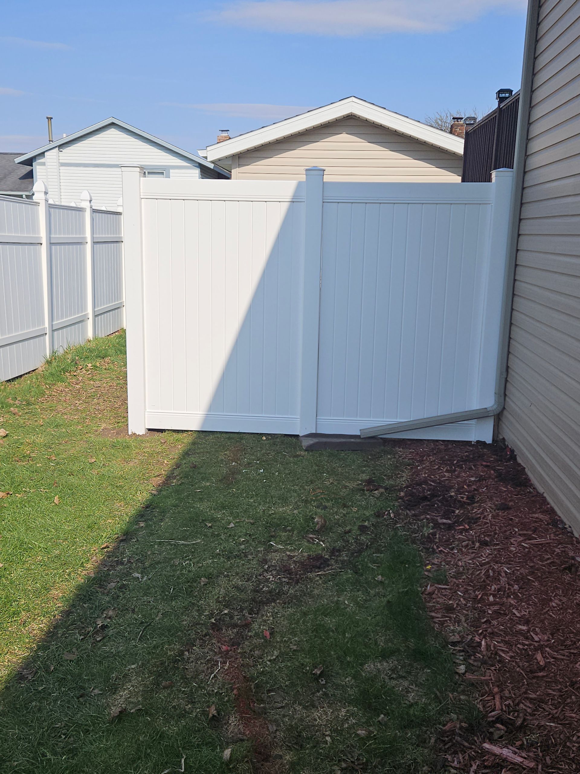 White vinyl gate and fence beside a narrow grass path between houses, under a blue sky.
