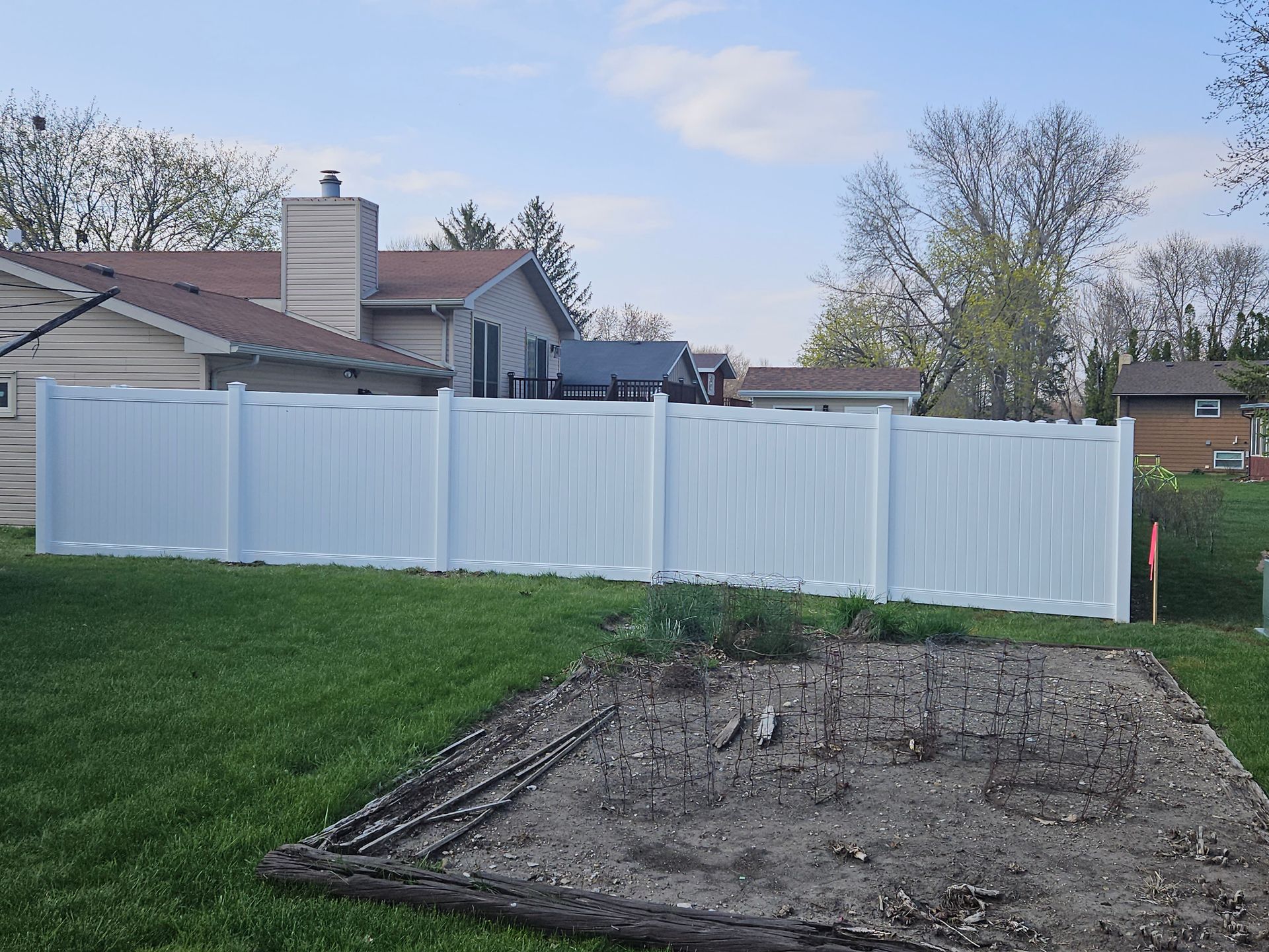 Backyard with white fence, grassy lawn, and a dirt patch beside a house at dusk