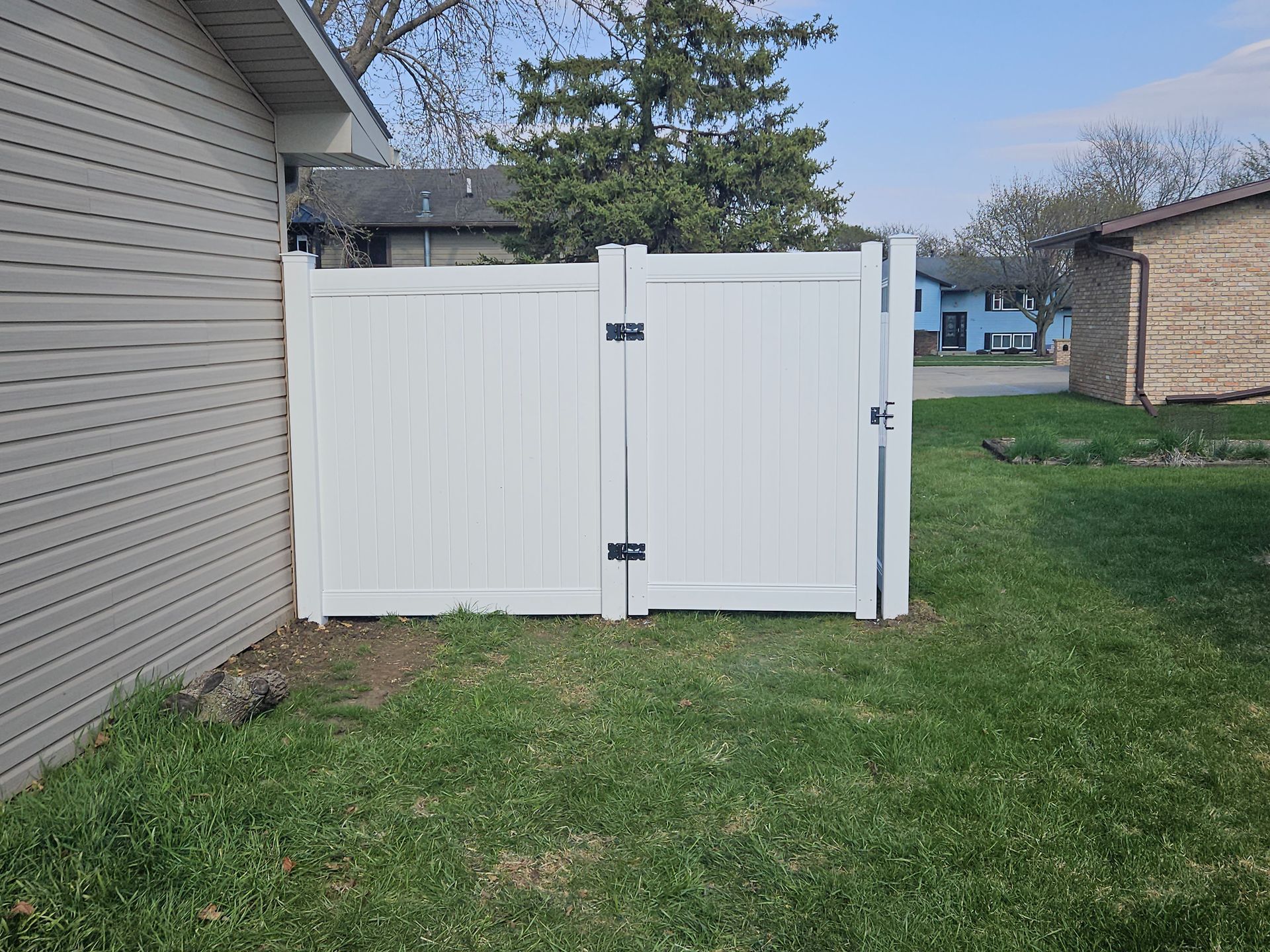White vinyl fence and gate beside a house, opening to a grassy backyard with trees and neighboring homes.