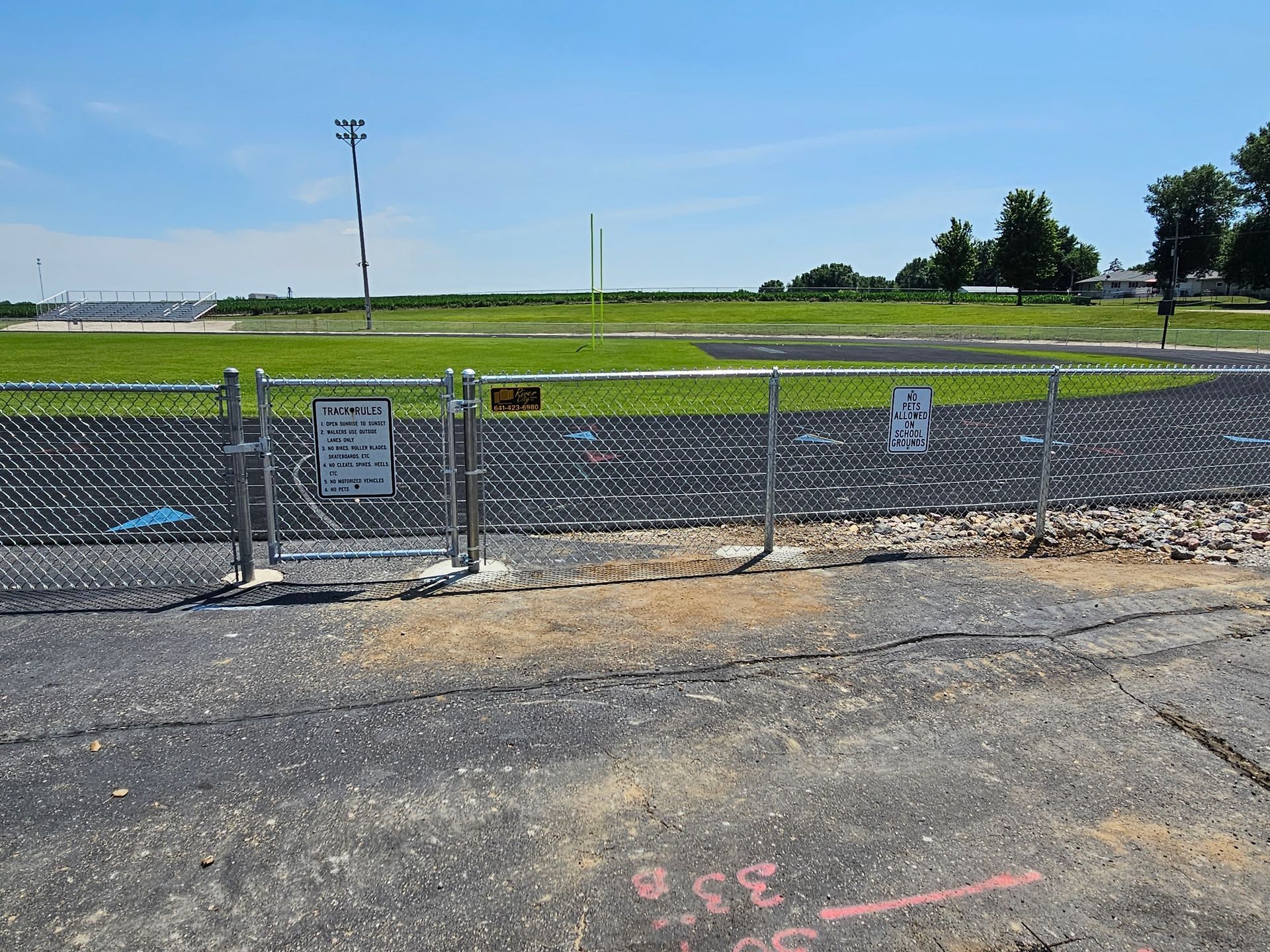 Chain-link fence around a grassy field under a clear blue sky, with gravel and pavement in the foreground.