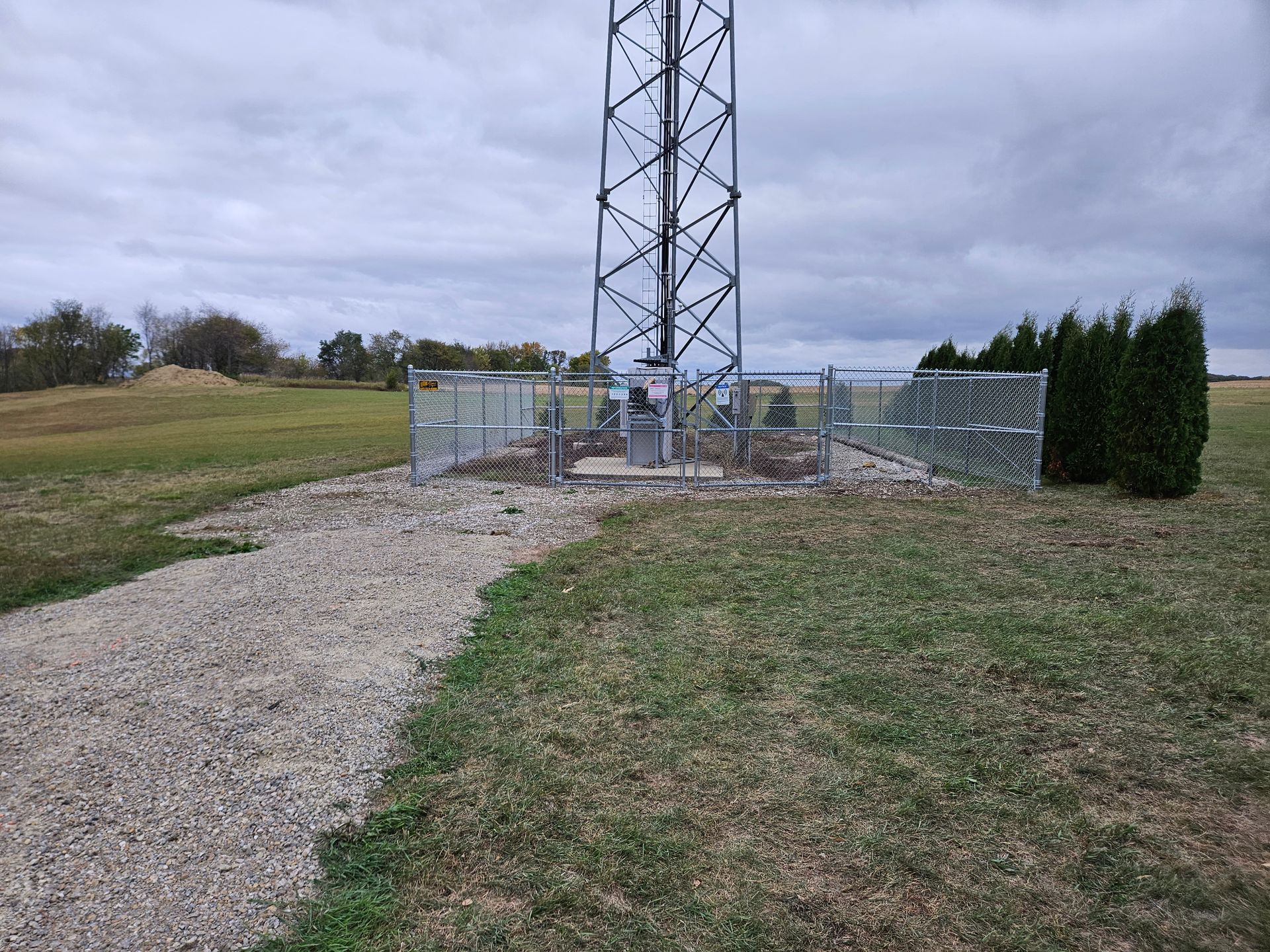 Gravel path leading to a fenced cell tower on a grassy hill under a cloudy sky