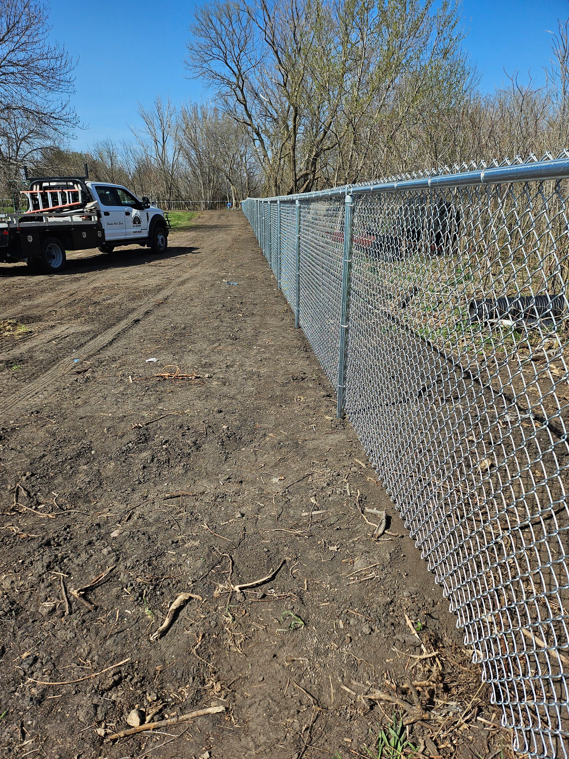 Chain-link fence along a dirt path beside a tow truck under trees on a sunny day