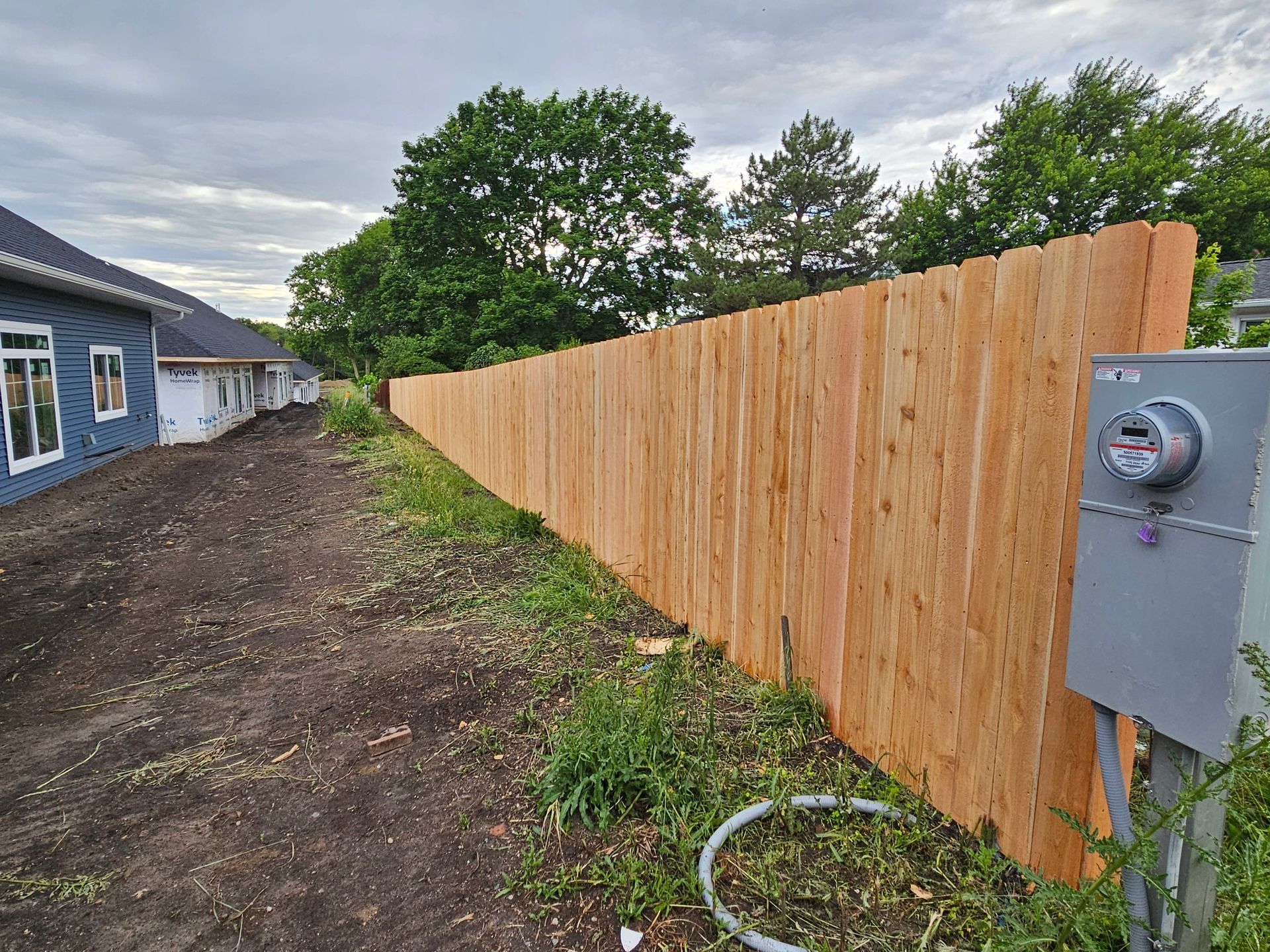 New wooden fence along dirt path beside blue house and utility meter under cloudy sky