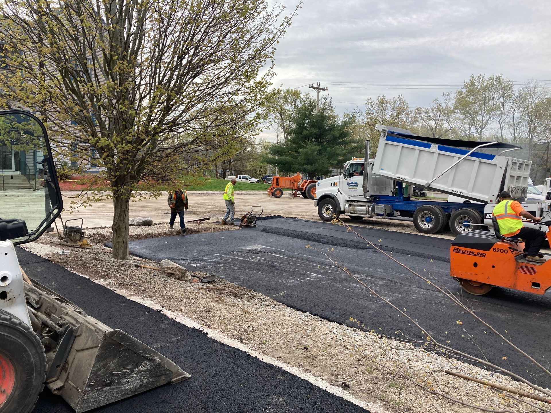 Asphalt paving: Dump truck unloading blacktop, workers, bobcat, and roller on a construction site.