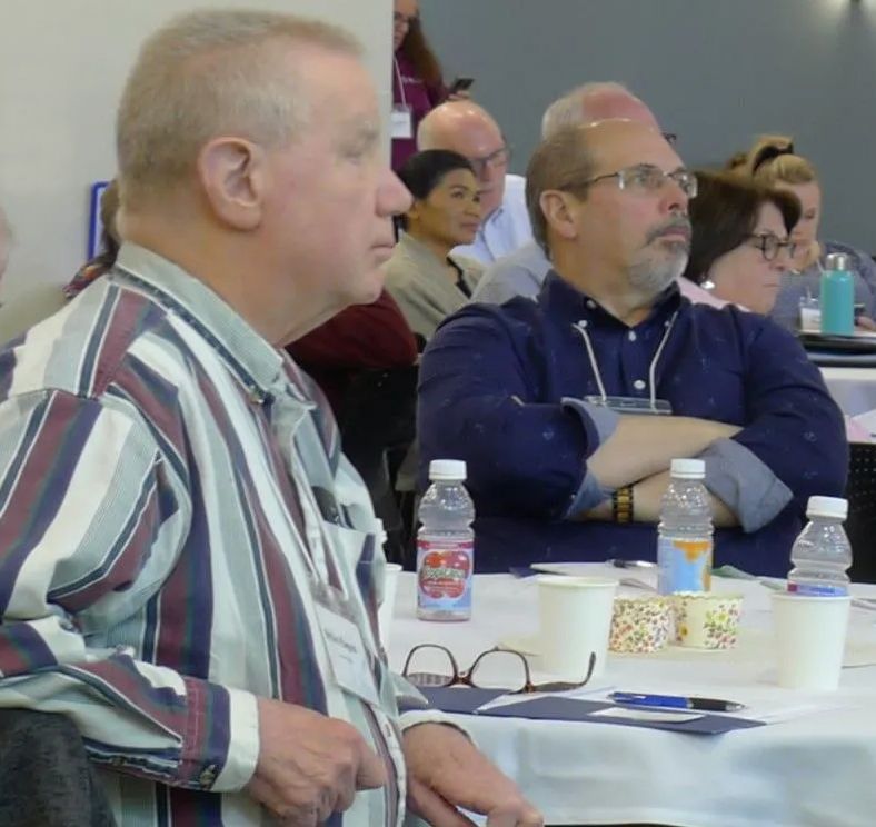 Two men listening at a conference table; other attendees in the background.